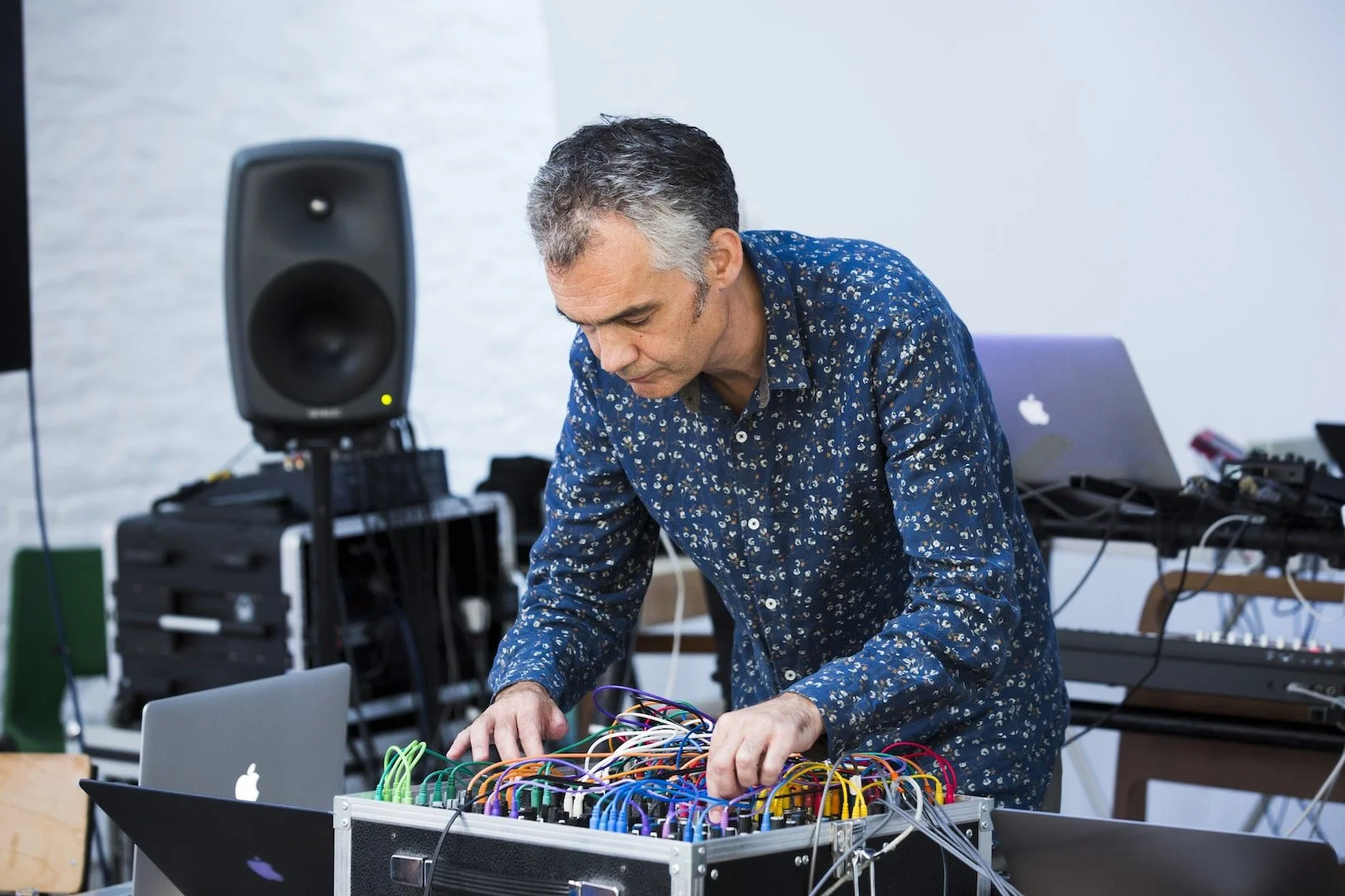 A man in a floral shirt working with electronic music equipment, including a patchbay with colorful cables, in a studio environment with a speaker, laptops, and other gear.