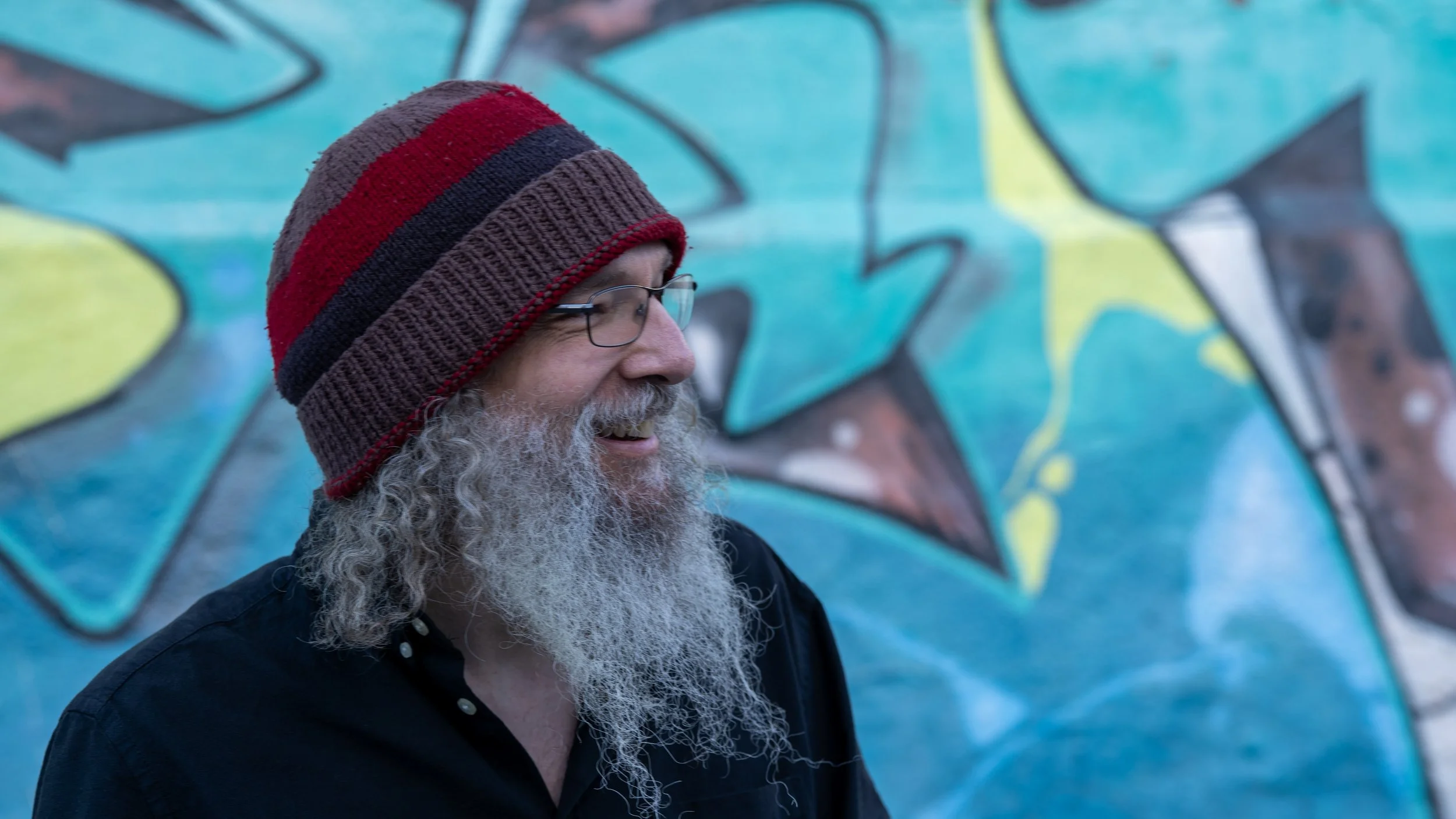 A man with glasses, a long gray beard, and a striped knit beanie hat is smiling while standing in front of a colorful graffiti wall.
