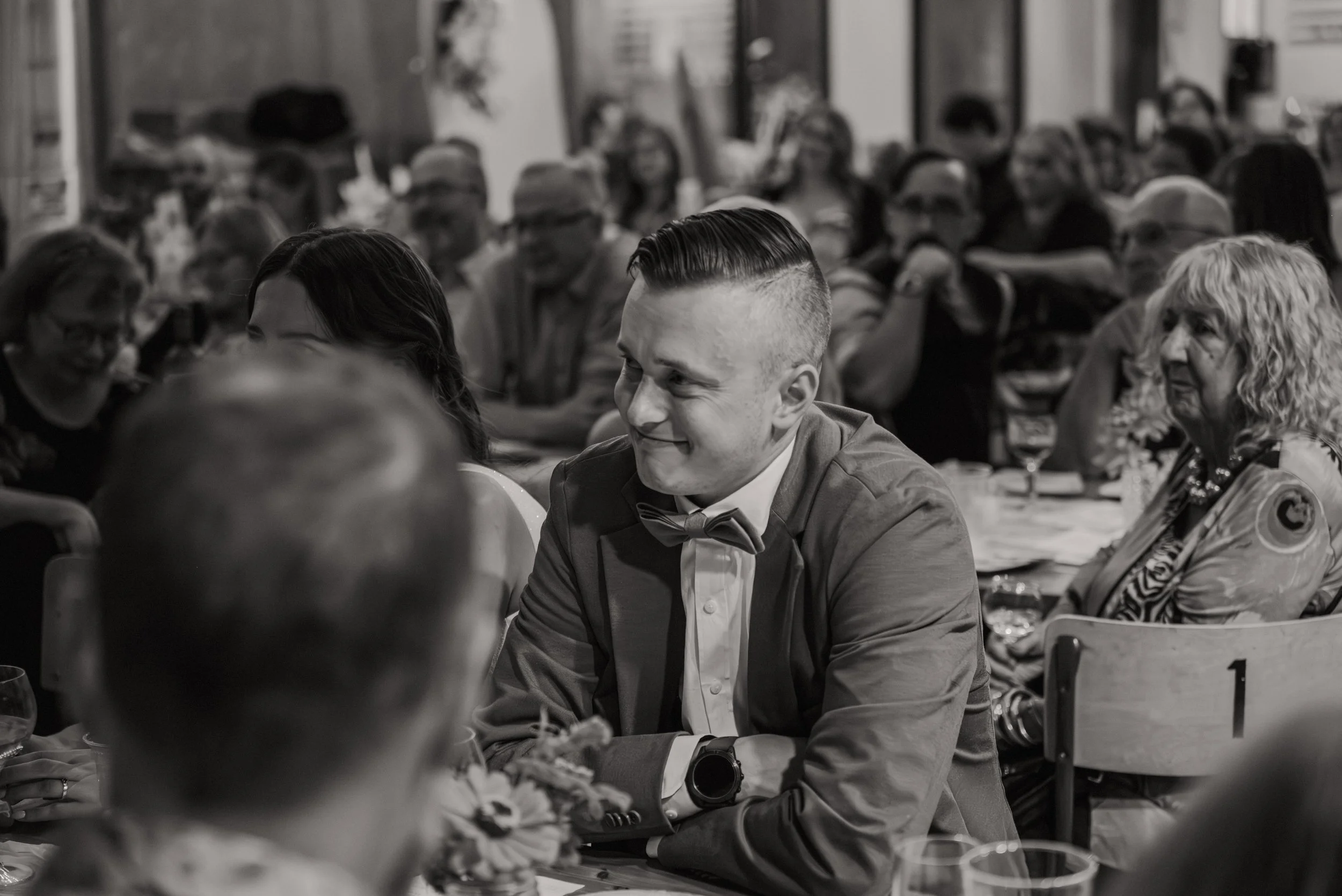 A young man with a slicked-back hairstyle, wearing a suit and bow tie, smiles while seated at a formal gathering or dinner event, surrounded by other guests in an indoor setting.