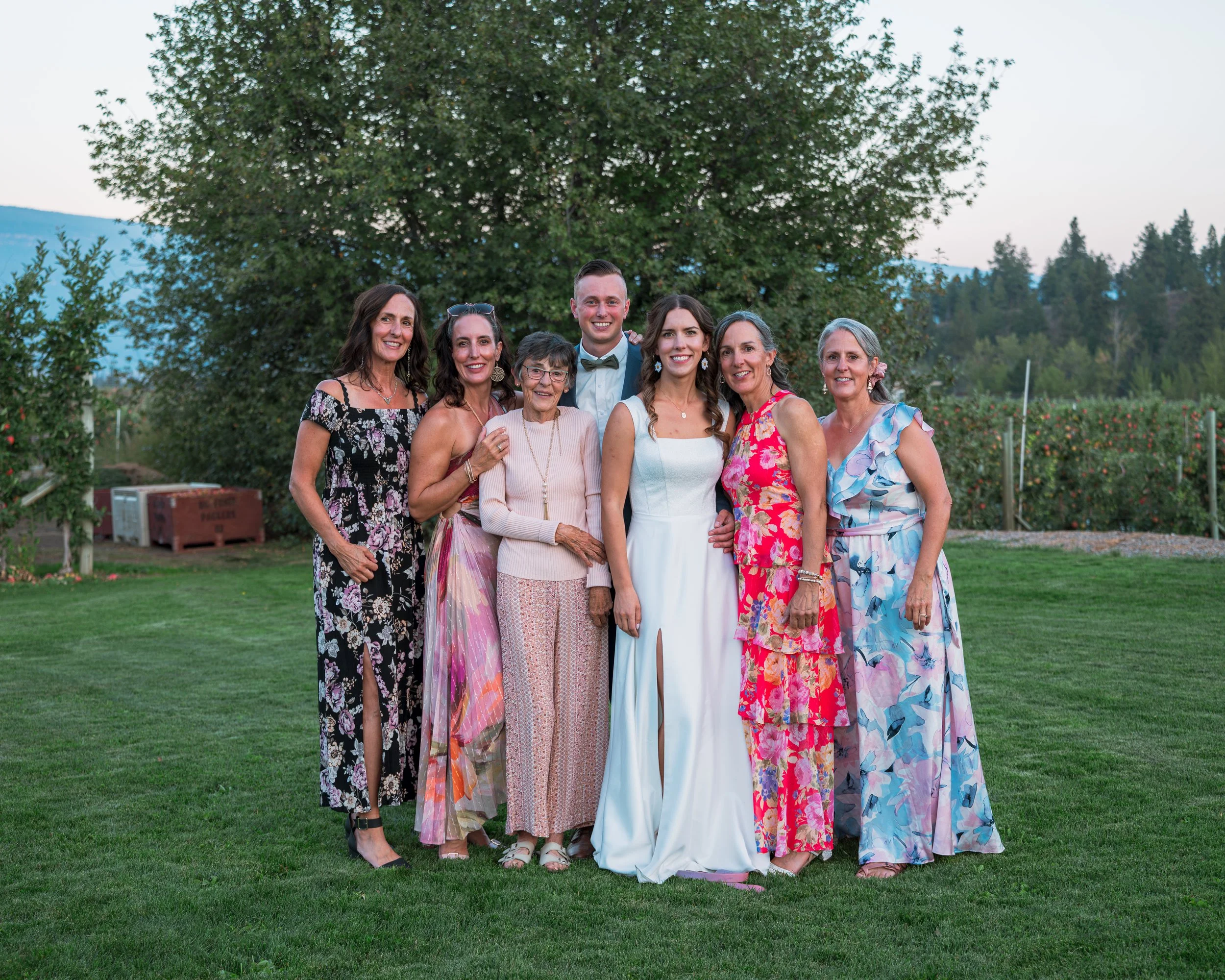 A group of eight women and one man, all dressed in semi-formal attire, standing on a grassy field outdoors with trees in the background. The man is in the center, wearing a light blue shirt with a bow tie, surrounded by women in colorful dresses.