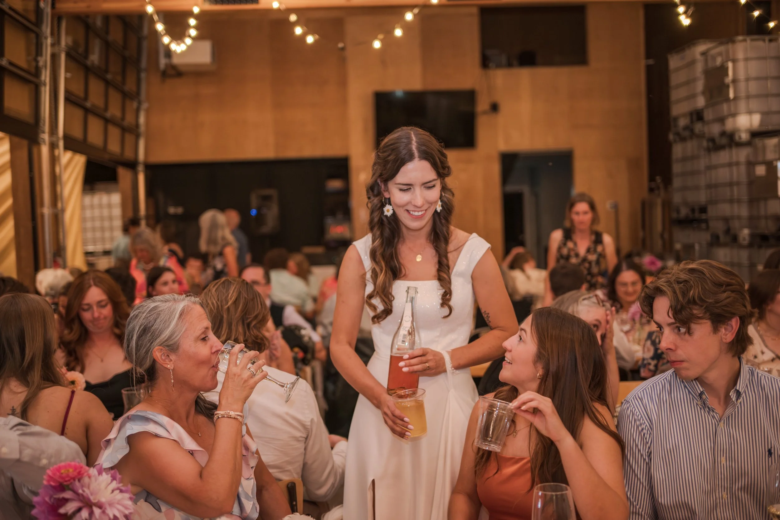 Women at a social gathering with drinks in a warmly lit indoor venue, some sitting and some standing, engaging in conversation.