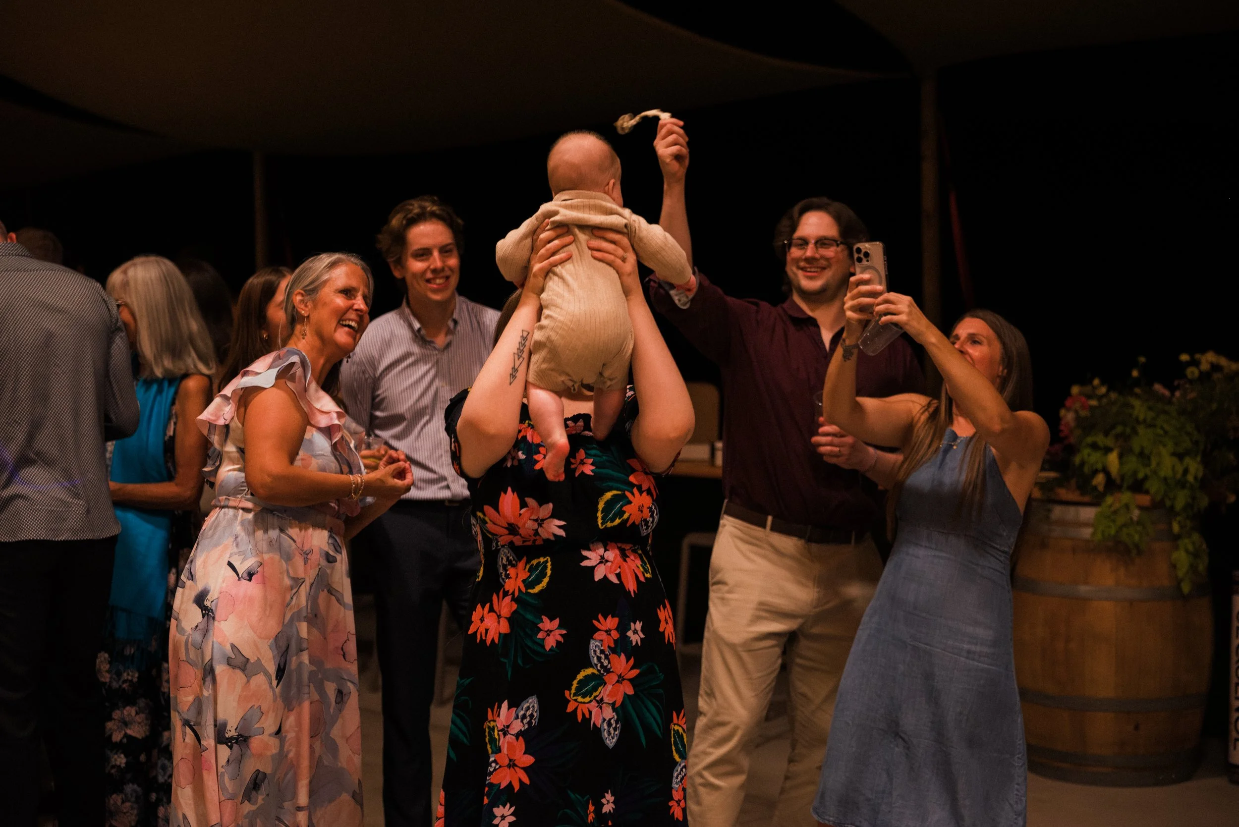 Group of people celebrating, with a woman holding a baby upside down and a man taking a photo at a party or gathering.