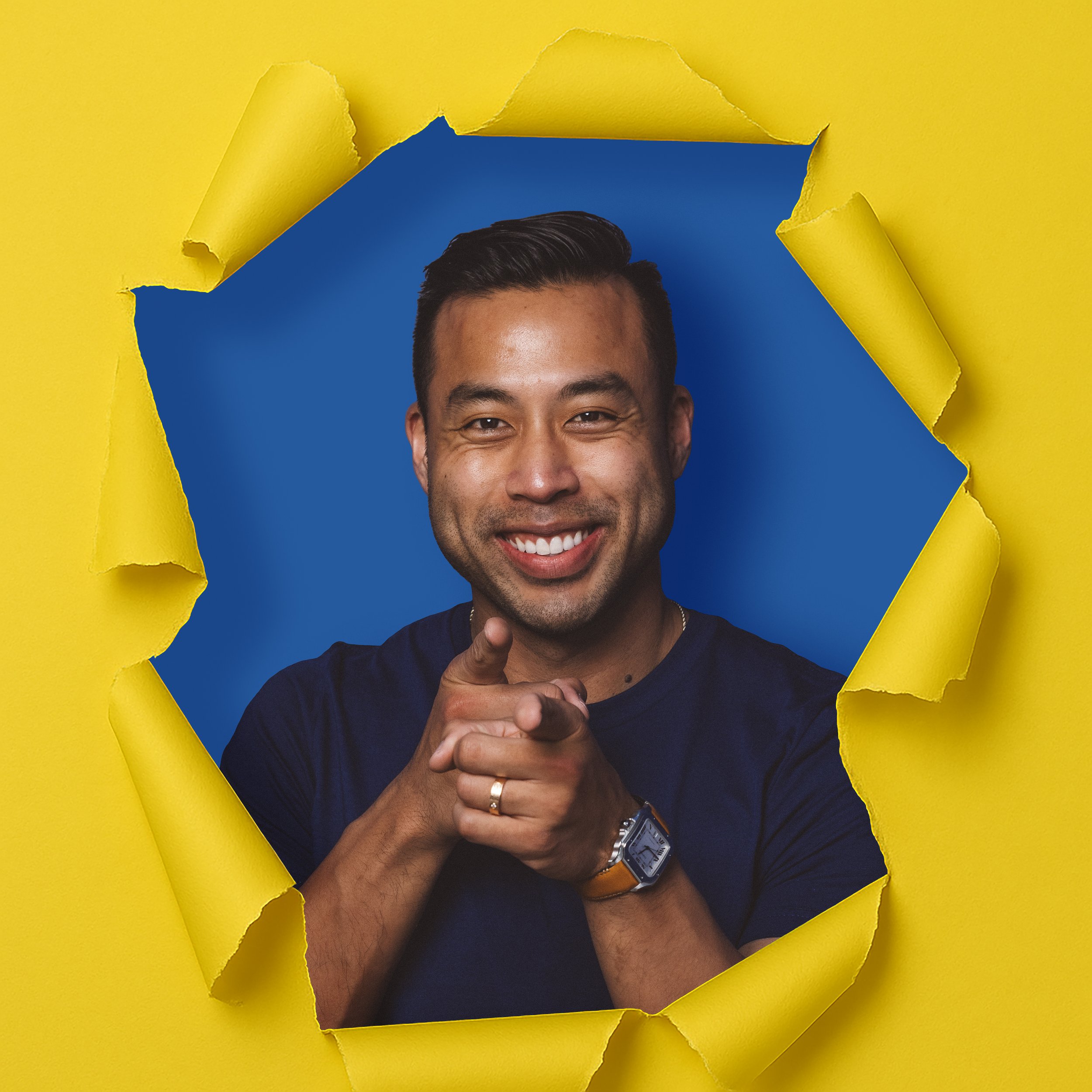 Man smiling and pointing at the camera through a torn yellow paper with a blue background.