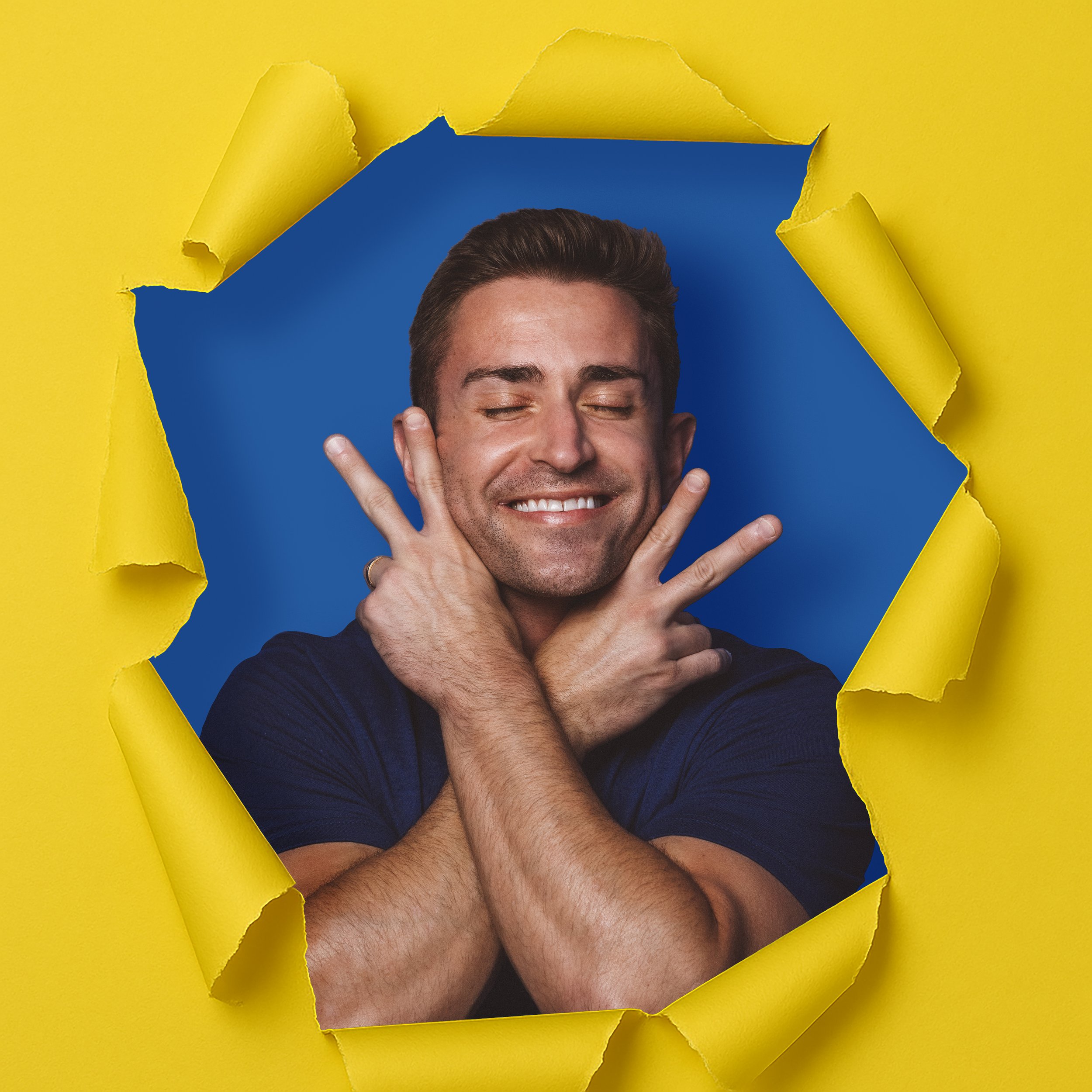 A man smiling with eyes closed, holding his hands to his face with index fingers and thumbs extended, looking happy, as if coming through a yellow paperhole with torn edges, revealing a blue background.