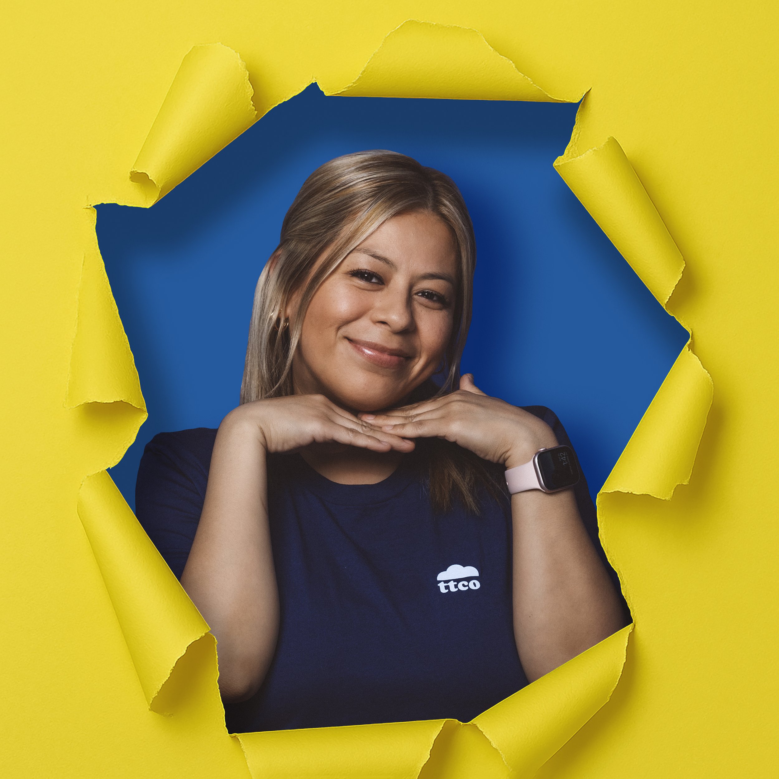 A smiling woman with blonde hair and a smartwatch posing through a torn yellow paper backdrop with a blue background.