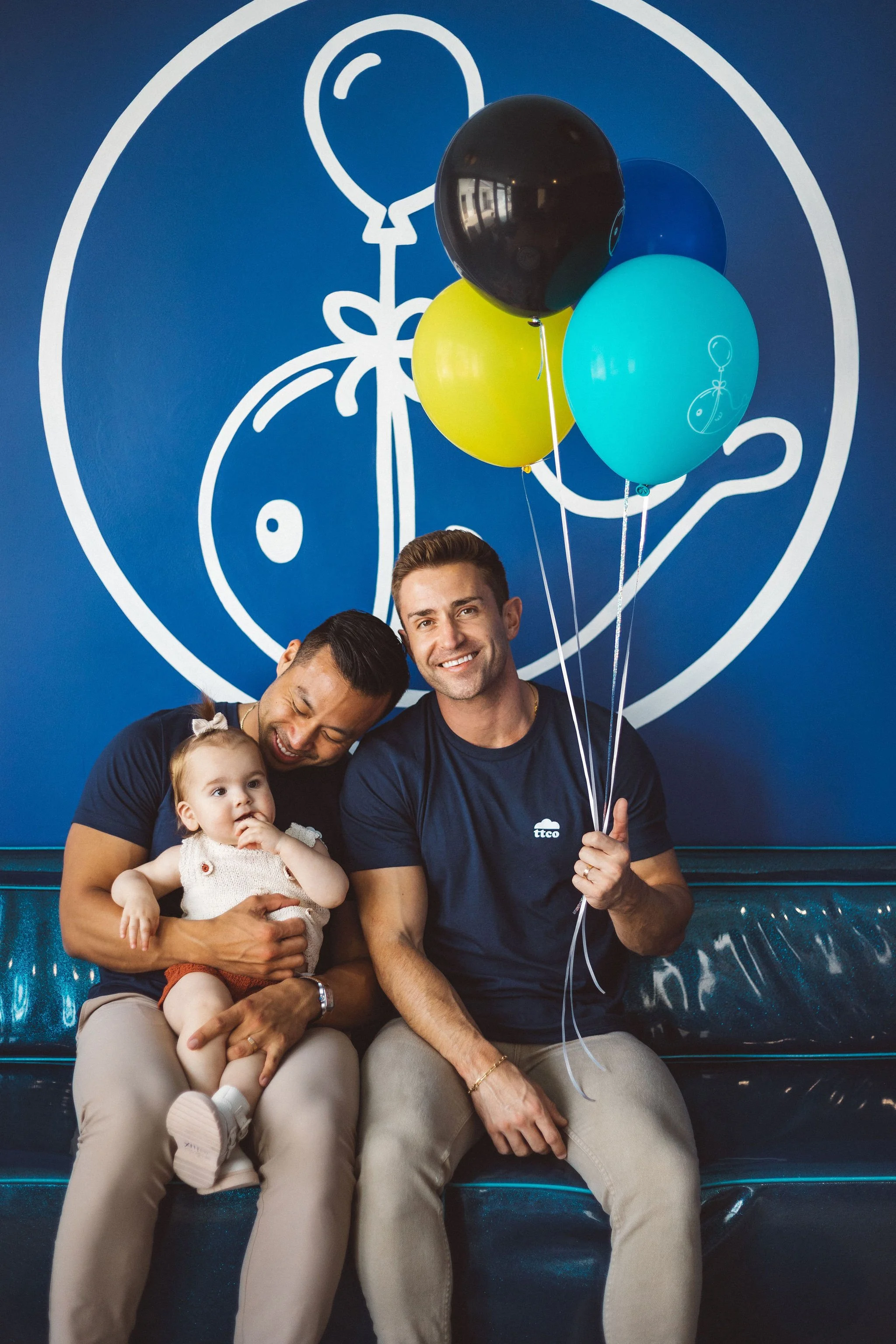 Four people sitting together in front of a blue wall with a white line drawing of a balloon and a tortoise, holding balloons, with a dark background reflection in the balloons.
