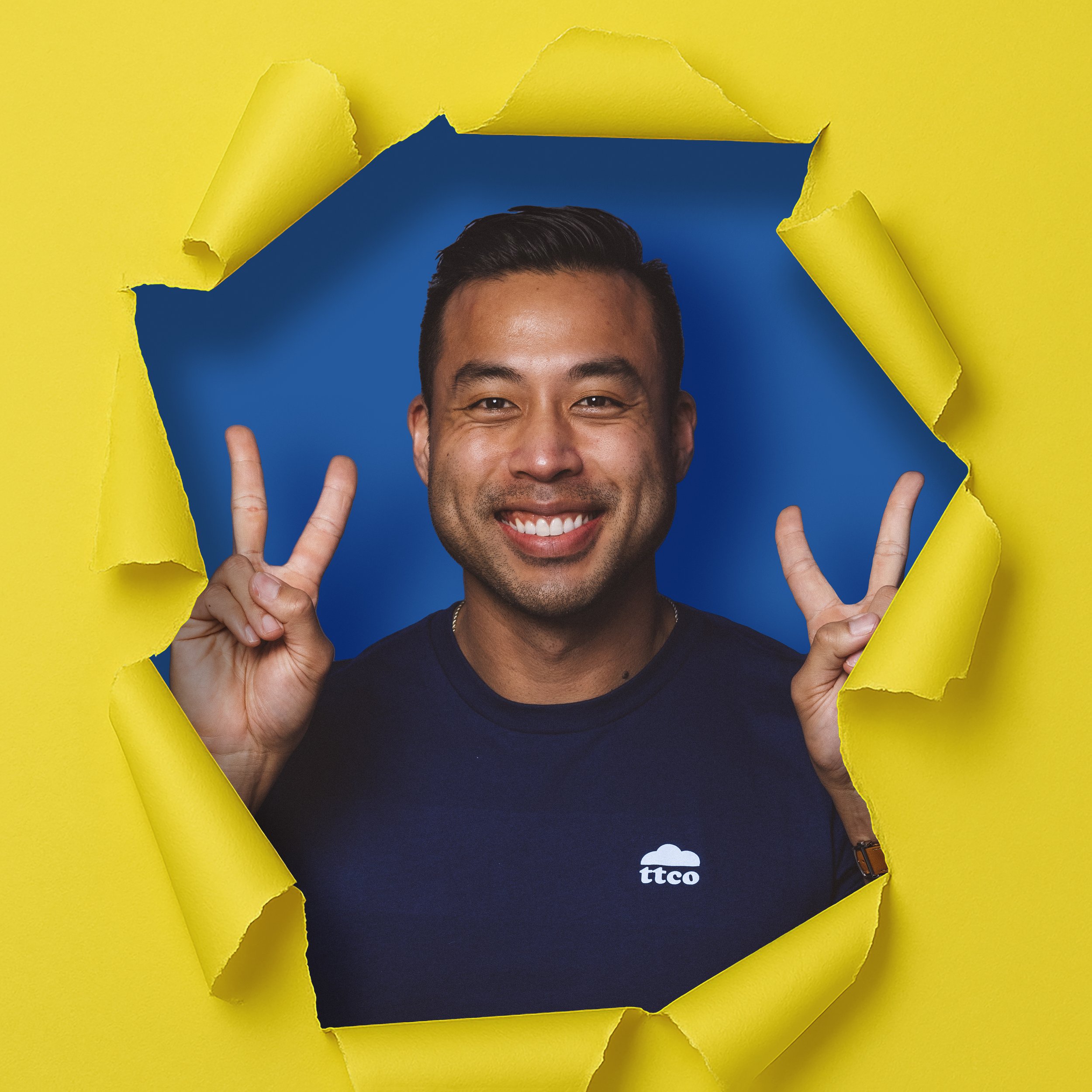 A smiling man showing two peace signs, peering through a torn yellow paper into a blue background.