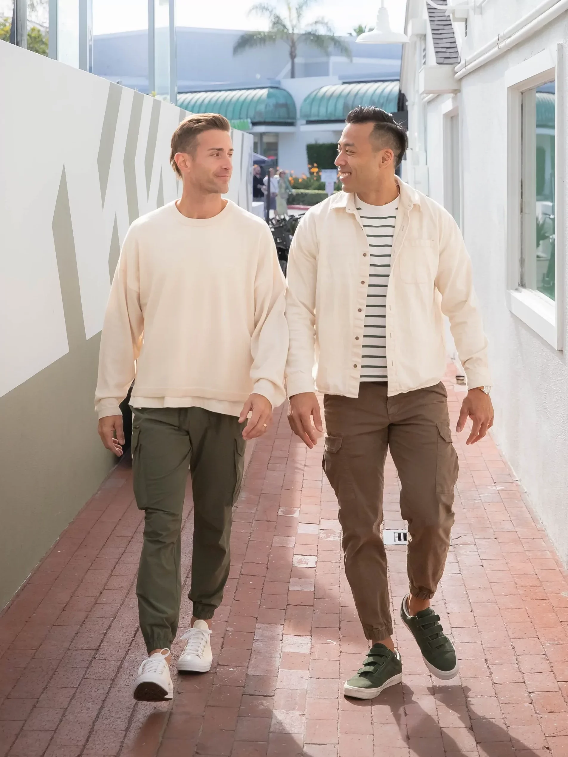 Two men walking together on a sidewalk, smiling and chatting, with a white building and a palm tree in the background.
