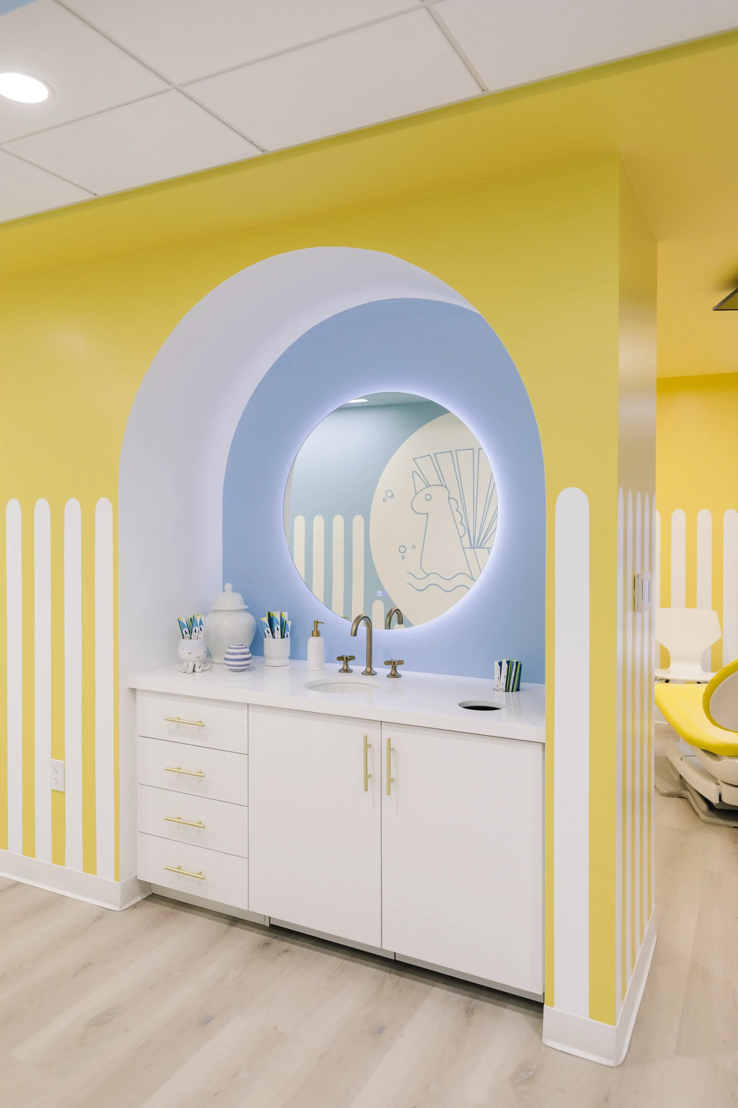 A modern bathroom with colorful yellow and white striped walls and a circular mirror. The white vanity has a round sink, gold handles, and various decorative items including containers with brushes and a white pot. There is a partial view of a yellow chair on the right and a blue wall with a chalkboard drawing of a unicorn with fins and bubbles.