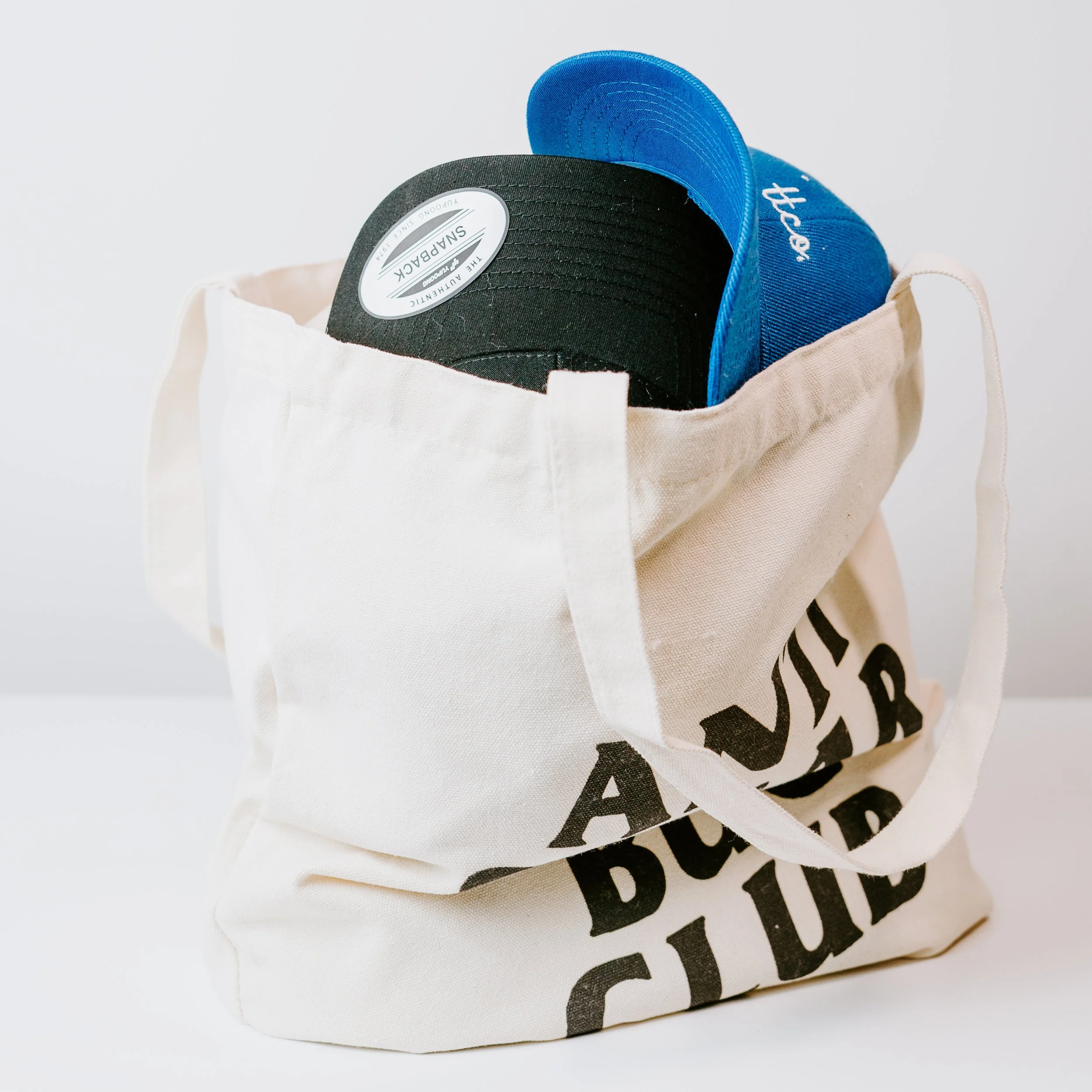 Close-up of two baseball caps, one black and one blue, peeking out of a white reusable bag with black text on it.