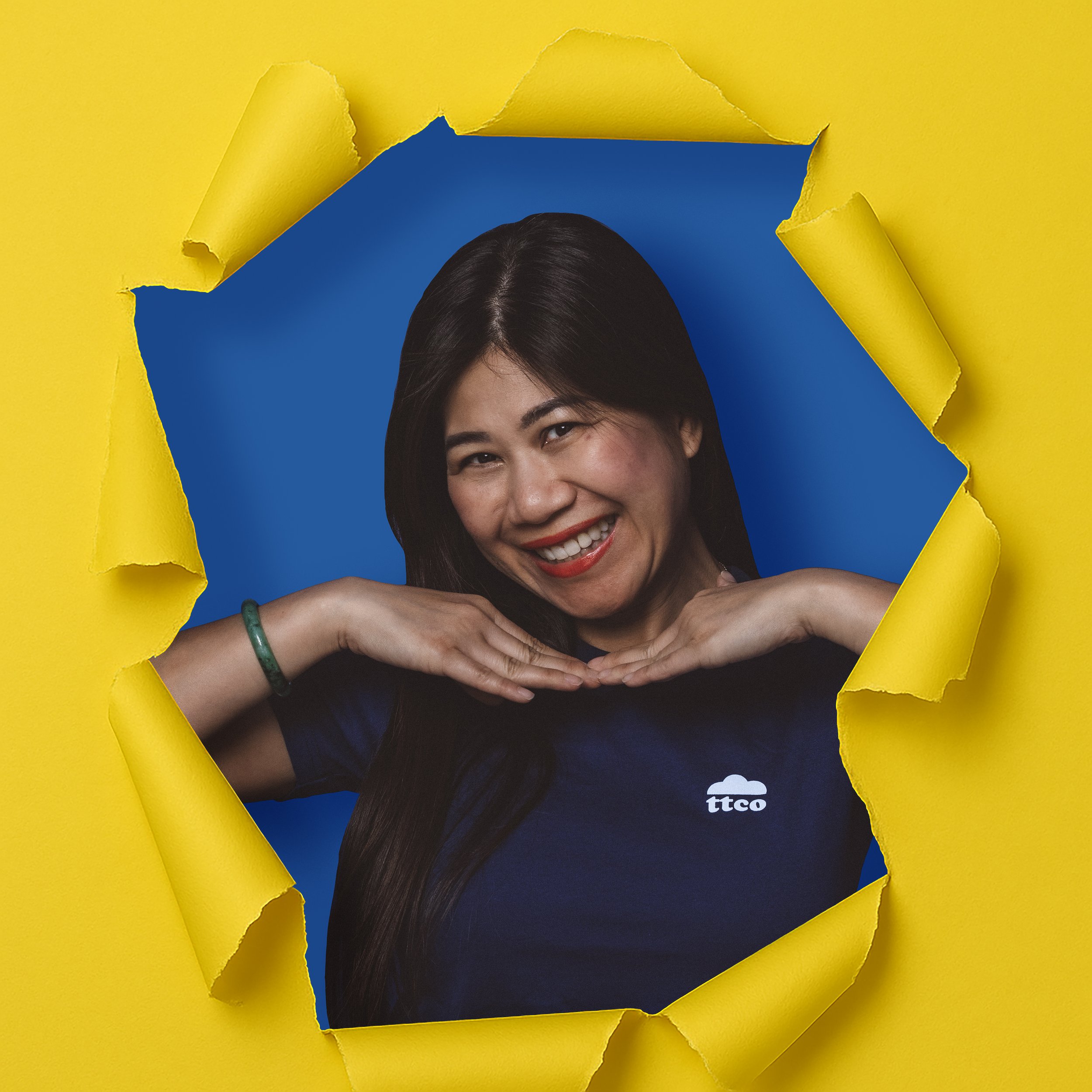 A woman with dark hair smiling and posing with her hands under her chin, seen through a torn yellow paper with a blue background. She is wearing a navy blue shirt with a logo and a green bracelet.