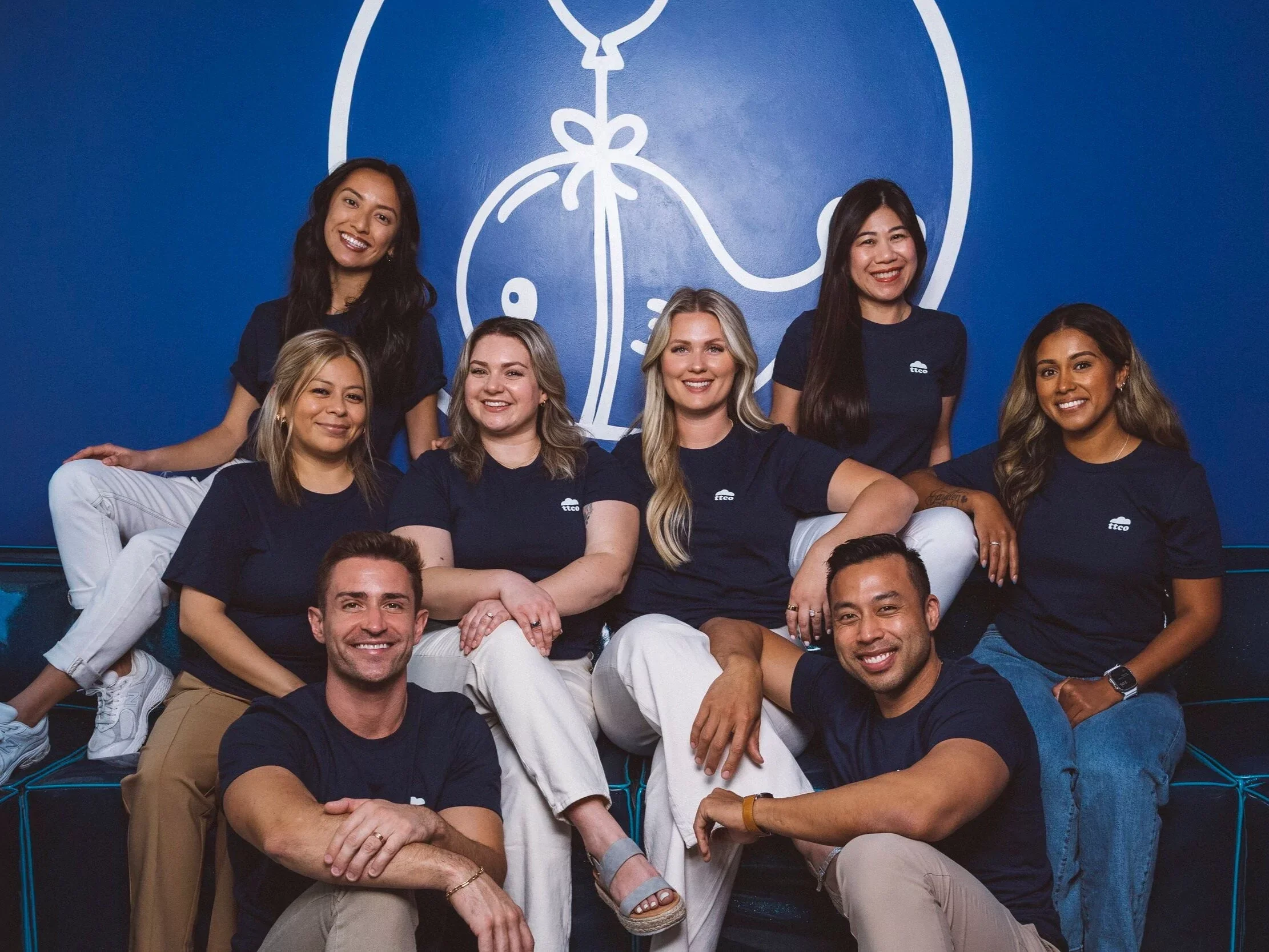 Group of diverse smiling people wearing navy blue T-shirts with a white logo, posed in front of a blue wall with a large white graphic of a pocket knife.