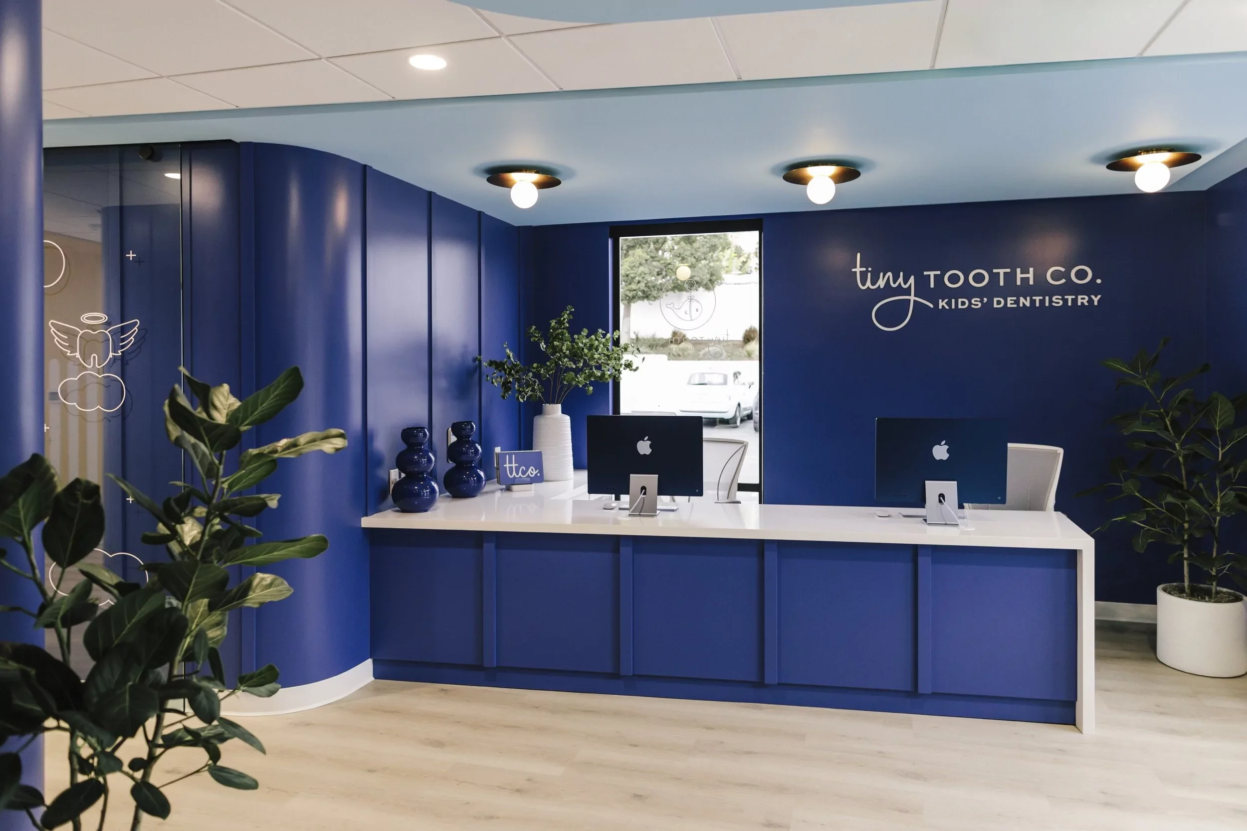 Reception area of Tiny Tooth Co. Kids' Dentistry with a blue wall, a white counter with computers and decorative vases, plants in white pots, and a window showing trees and parked cars outside.