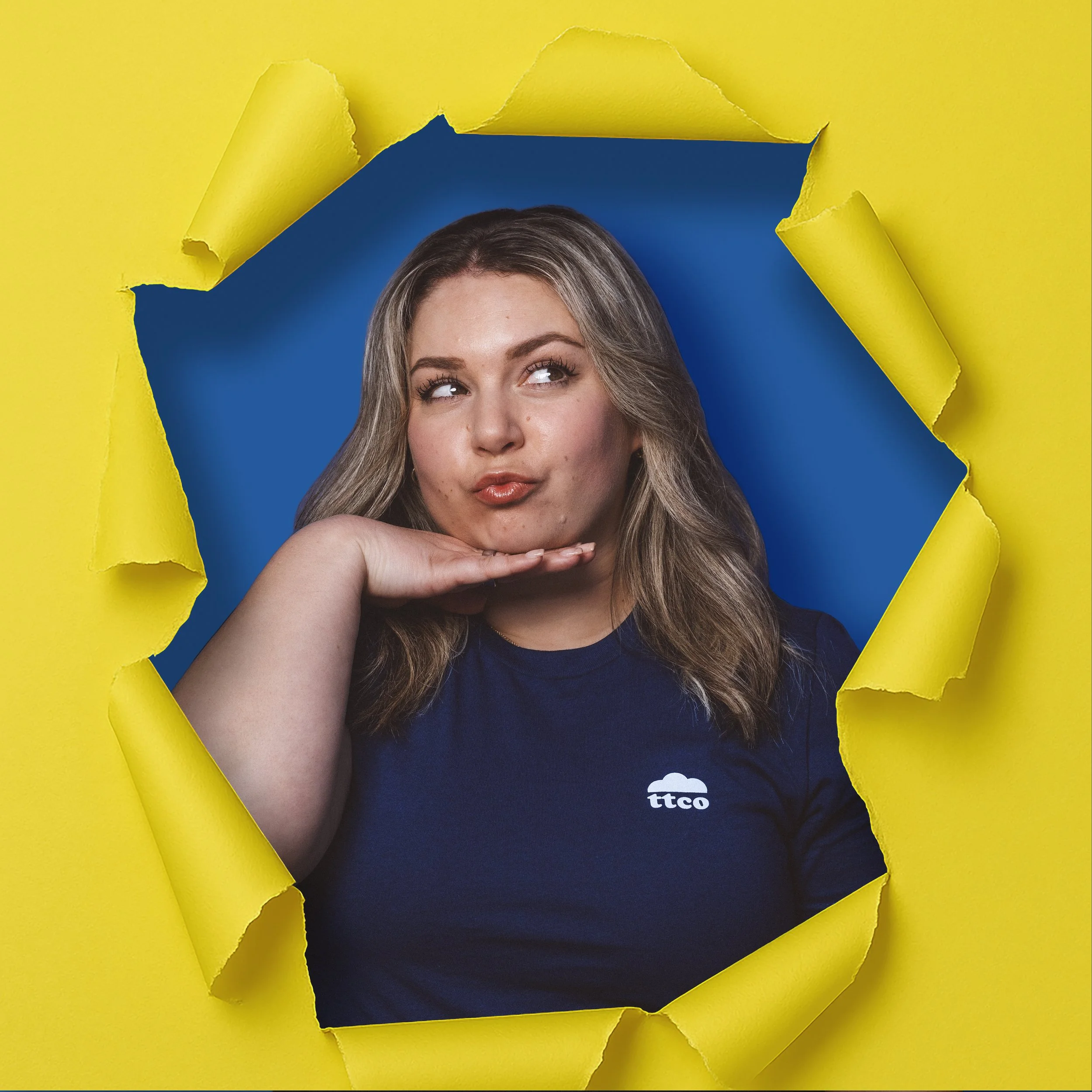 A woman with long wavy hair making a playful face, resting her chin on her hand, seen through a torn yellow paper backdrop.