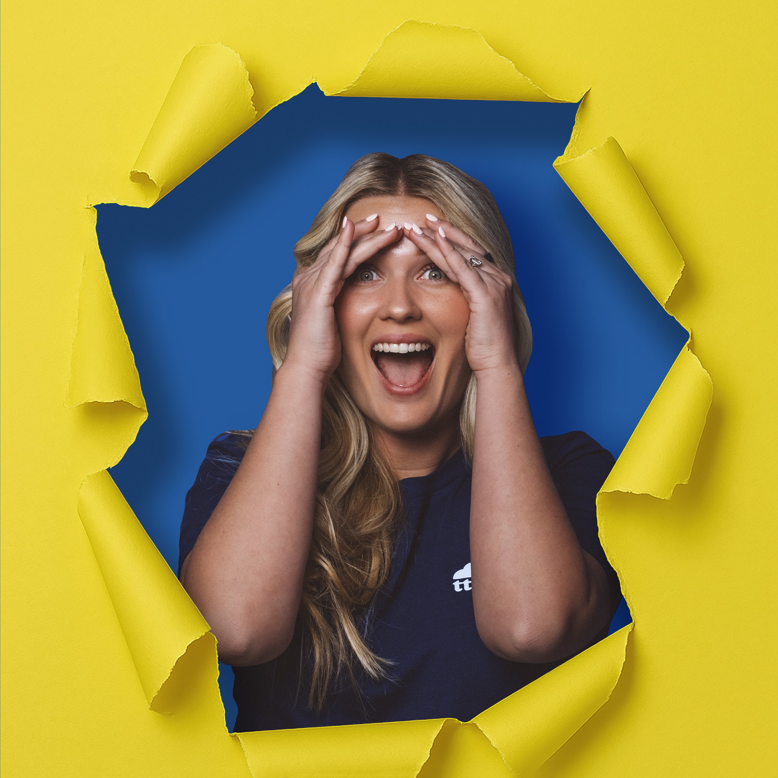 A woman with long blonde hair, wearing a dark blue shirt, looks excited as she peeks through a torn paper hole in a yellow background, with her hands on her forehead and mouth open in a joyful expression.