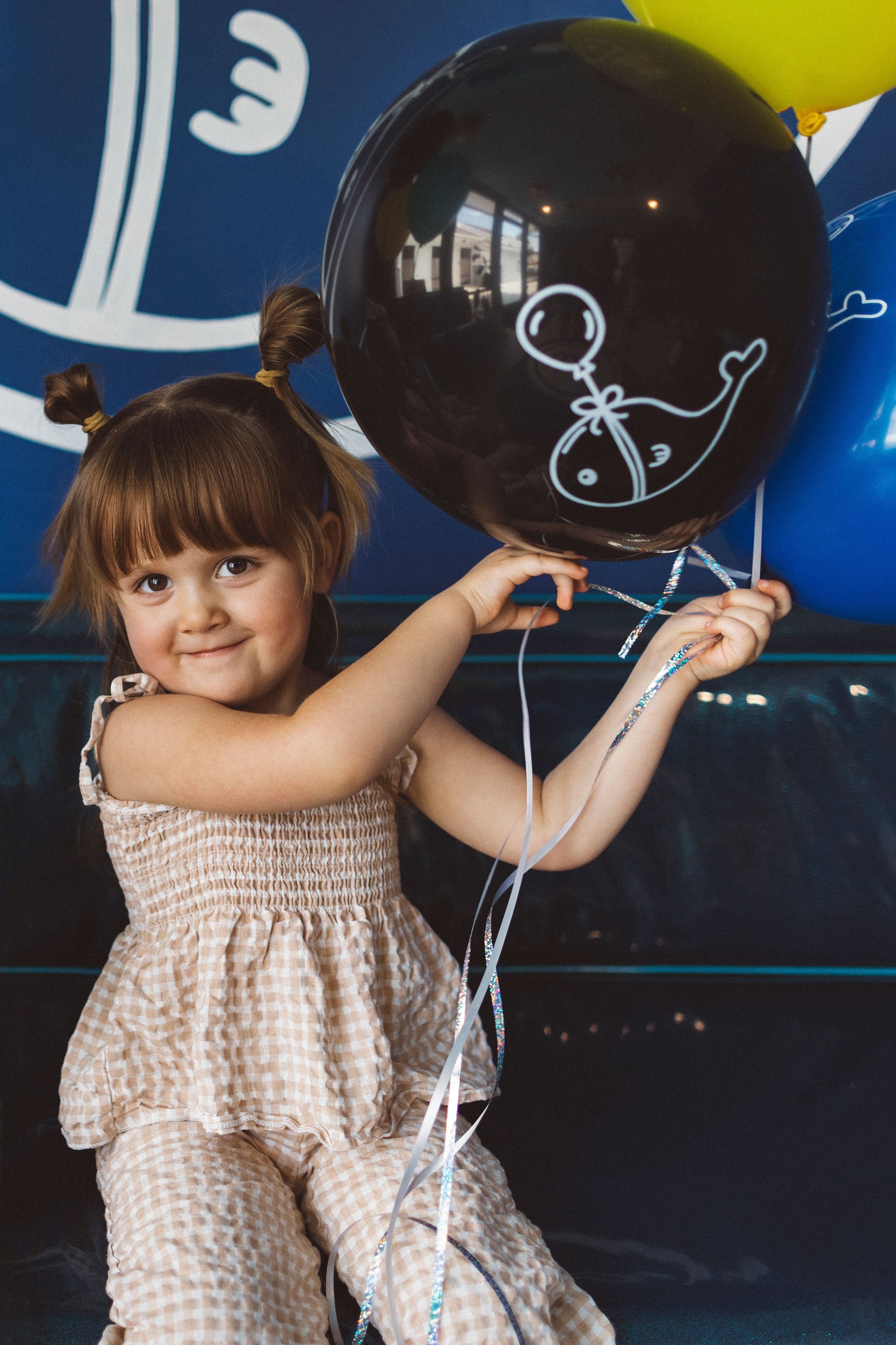 A young girl with brown hair styled in two pigtails, wearing a light-colored checkered dress, holding black and blue balloons with white balloon illustrations, standing in front of a dark background and a blue wall with a white logo.