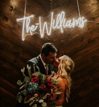 A couple kisses in front of a custom last-name sign while the bride holds a cascasding and gorgeous bouquet of fresh flowers at their lavish wedding in Fort Wayne, IN. Details and day planned by bella and fern event planning.