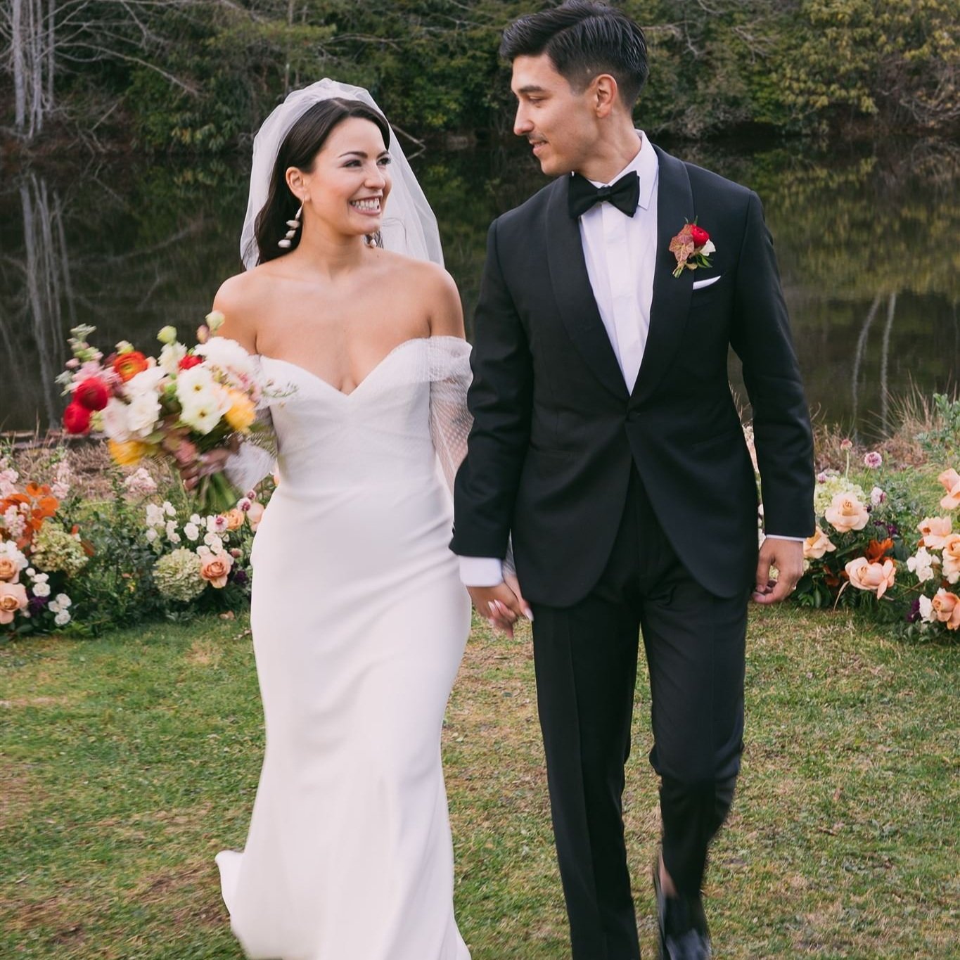 A bride and groom holding hands and smiling outdoors, with a background of flowers and trees.