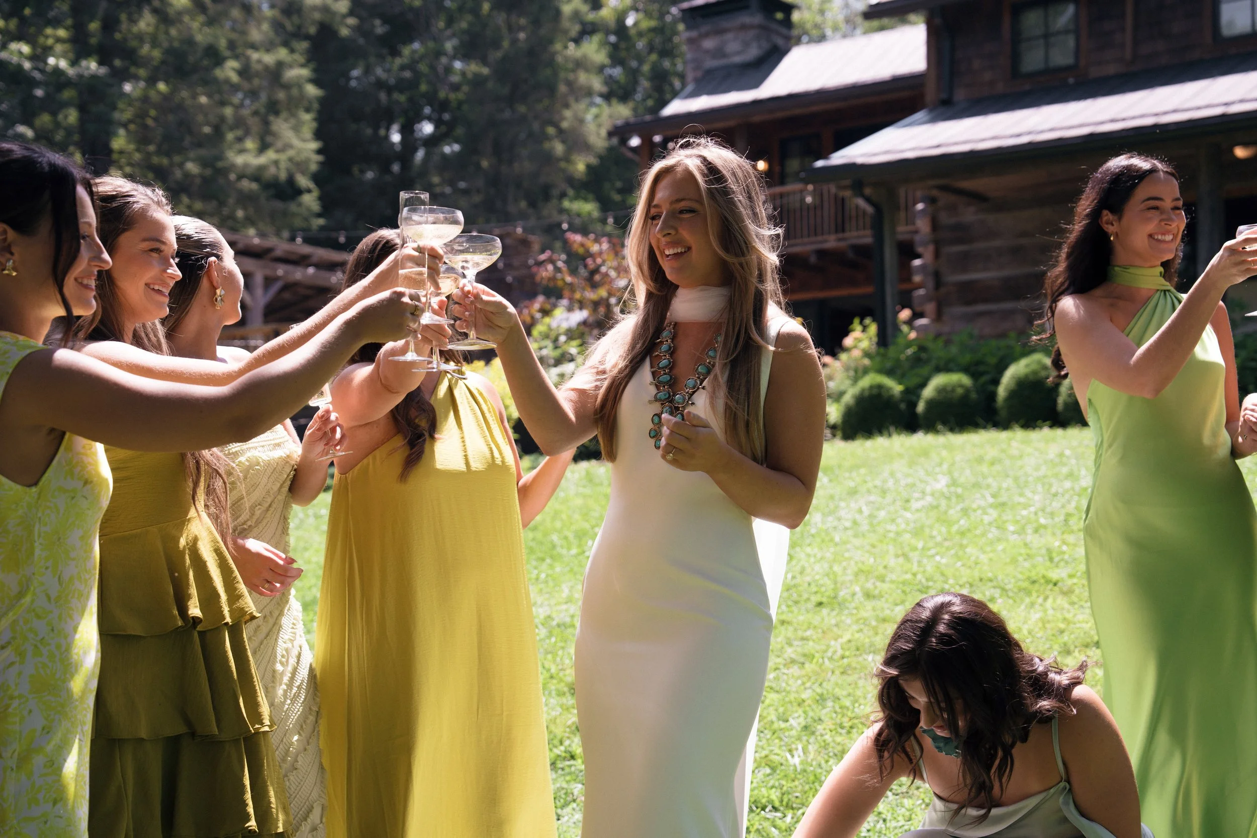 Group of women celebrating outdoors, raising glasses of champagne or sparkling wine, with a house and trees in the background, appears to be a sunny day.