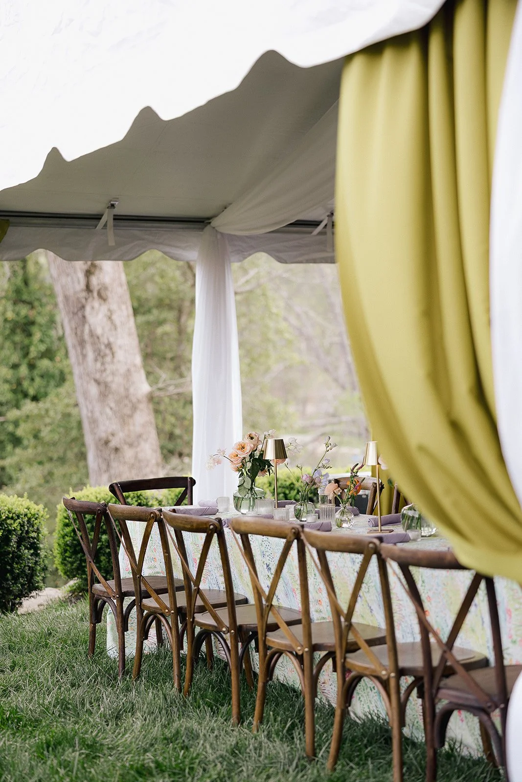 Outdoor decorated table set under a white tent with yellow curtains, featuring a floral tablecloth, flower arrangements, and small lamps, surrounded by wooden chairs on a grassy area with trees in the background.