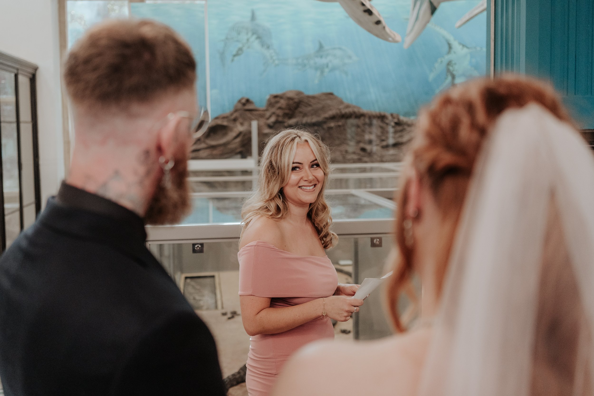 A wedding ceremony with a smiling woman in a pink dress holding papers, standing in front of an aquarium with a bride and groom partially visible.