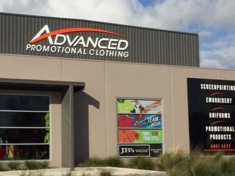 Exterior of a building with a sign that reads 'Advanced Promotional Clothing' and a window display advertising screen printing, embroidery, uniforms, promotional products, and a phone number.