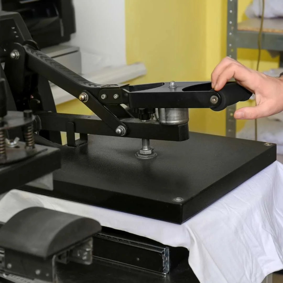 A person's hand is pressing a button on a large heat press machine used for transferring designs onto fabric, with a white garment underneath.