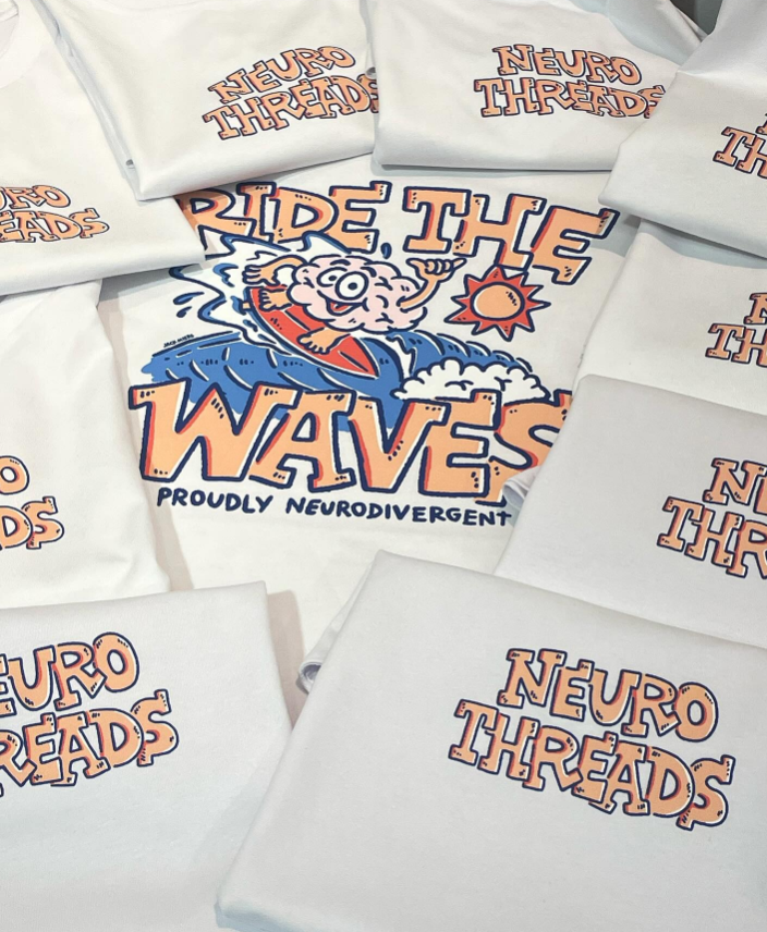 Multiple white t-shirts with logos. One shirt in the center features a cartoon brain riding a surfboard, with the text "Ride the Waves" and "Proudly Neurodivergent." The other shirts display the "Neuro Threads" logo.