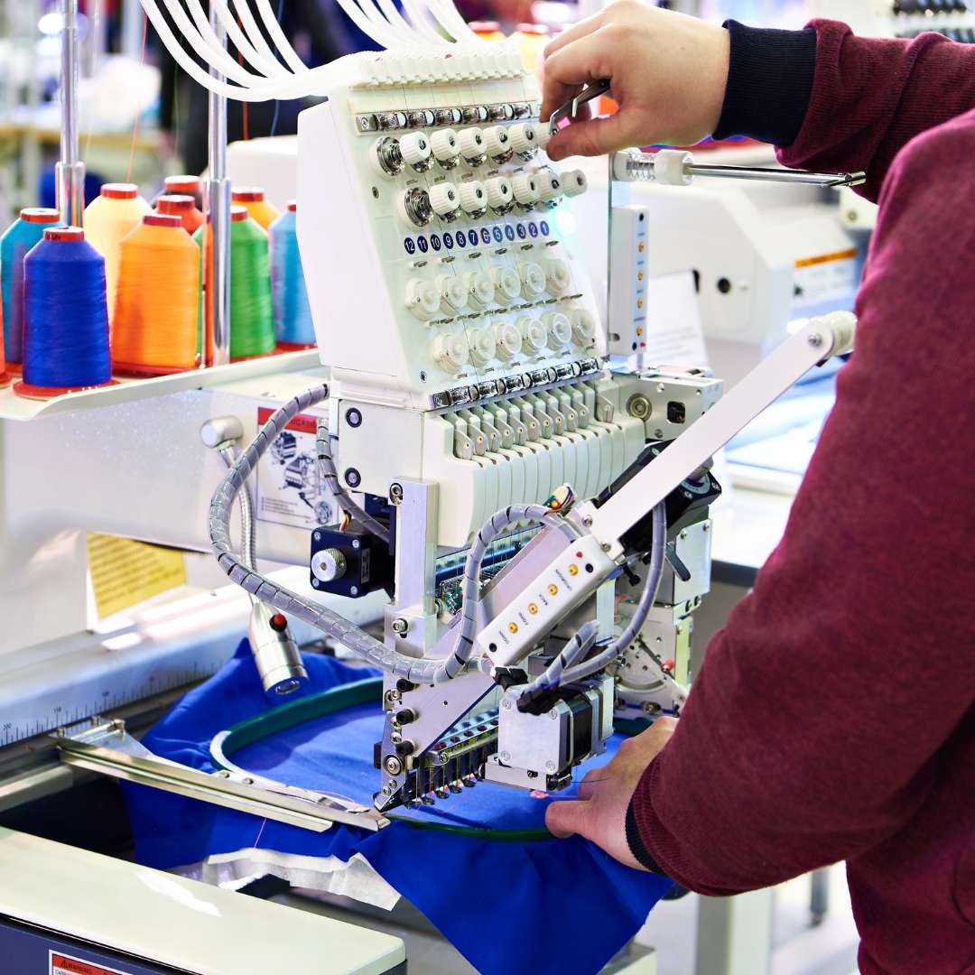 A person operates a sewing machine for embroidery or stitching on blue fabric in a workspace, with spools of colorful thread in the background.