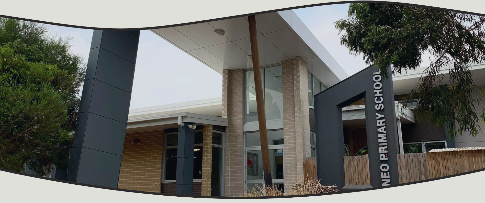 Exterior of Neo Primary School with modern architecture, brick and glass windows, and a sign reading 'NEO PRIMARY SCHOOL' near a wooden fence and surrounded by trees.