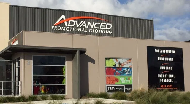 Exterior of a commercial building with signs for Advanced Promotional Clothing and JB's Wear, advertising screen printing, embroidery, uniforms, and promotional products