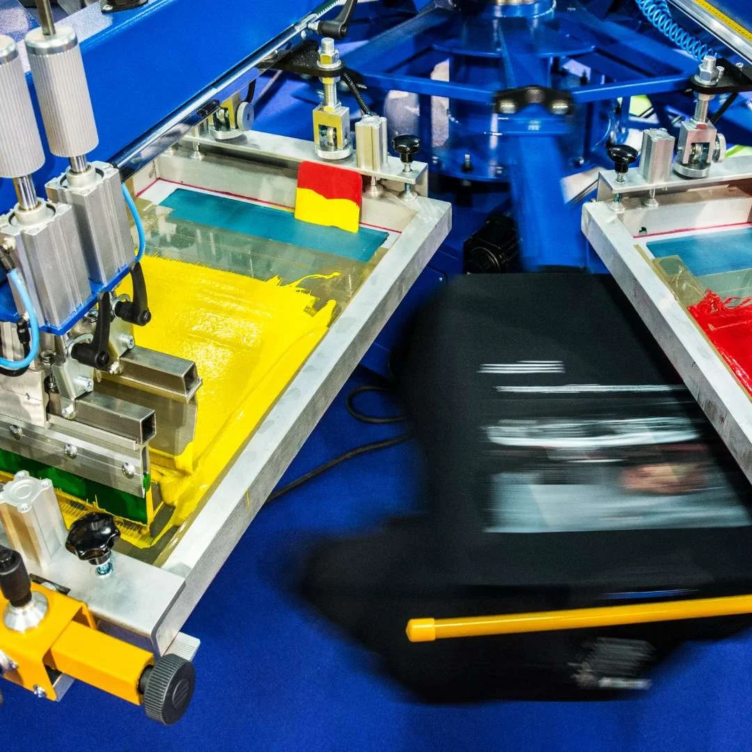 Automated screen printing machine with yellow and red paint on a glass surface, in a manufacturing setting with blue machinery in the background.