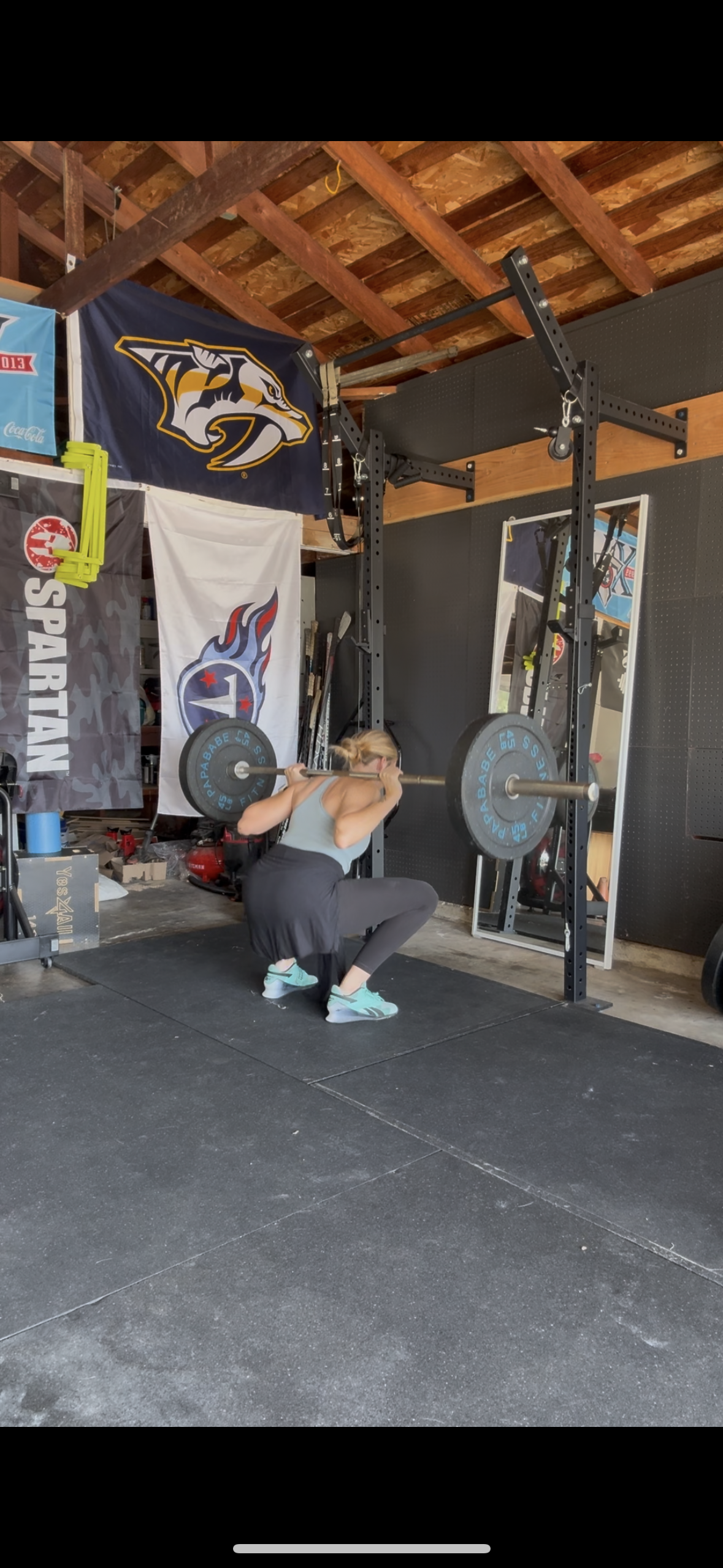 A woman in a gym is performing a squat while holding a barbell on her shoulders. She is centered in front of a black power rack, wearing a light blue workout top and black leggings. The gym has black rubber flooring, and the background features flags with logos and a mirror, with an exposed wooden roof.