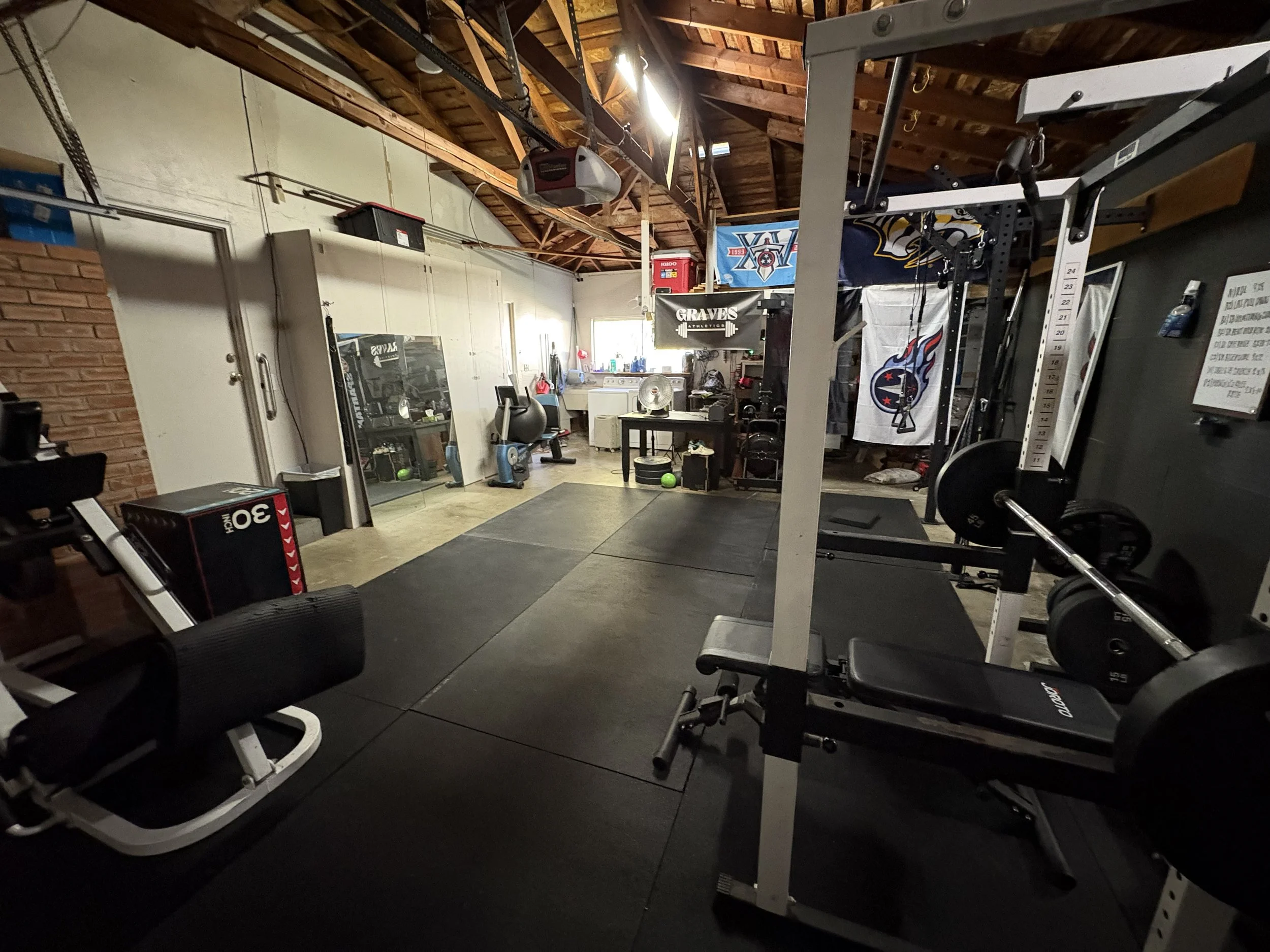 A home gym with weightlifting equipment, a bench press, and a black rubber flooring. The background shows a work area with various fitness accessories, banners with sports team logos, and a large open space with a high wooden ceiling.