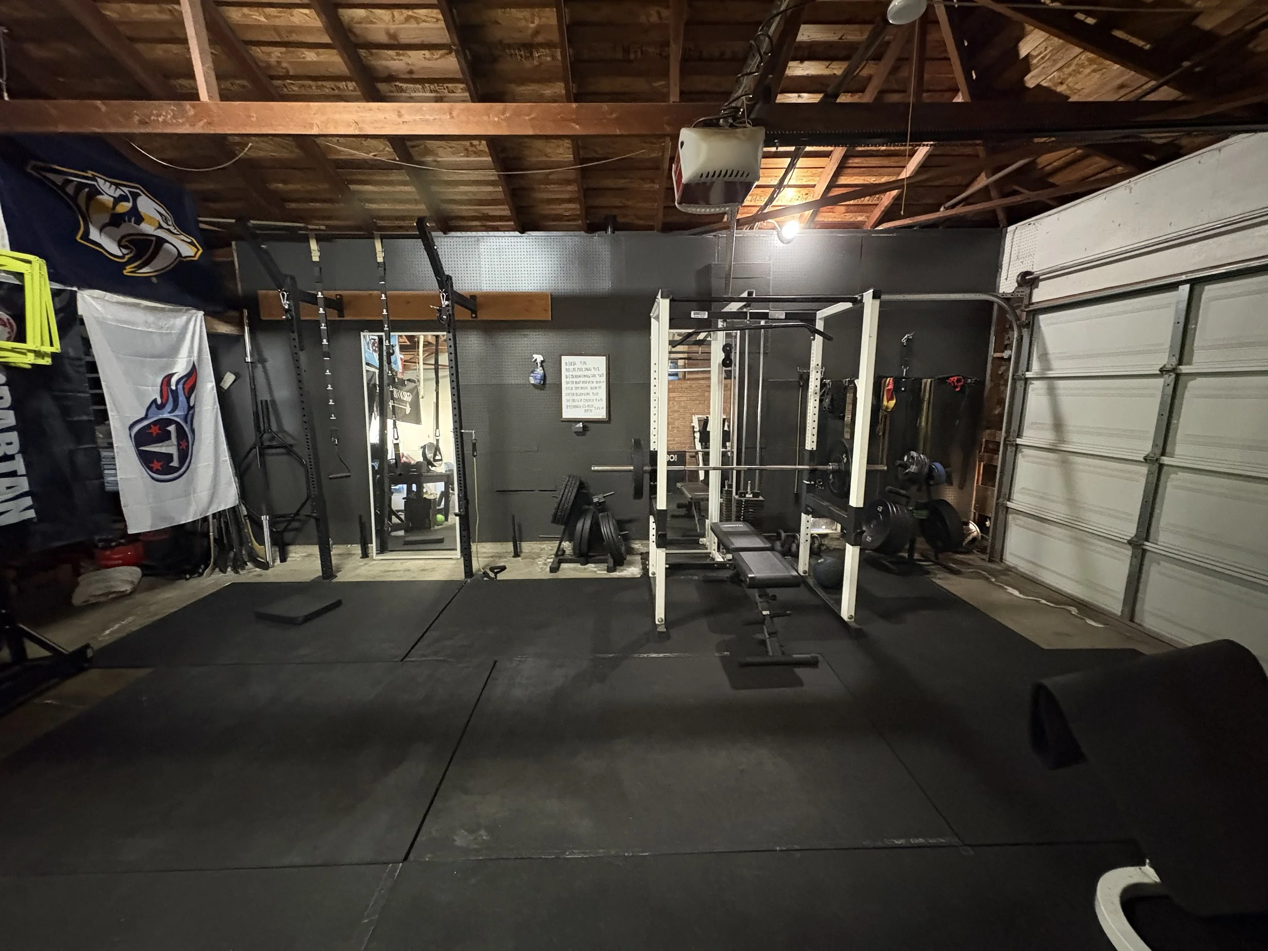 Home gym with workout equipment, weight rack, barbells, weight plates, and competition flags, in a garage with a garage door on the right and a wooden ceiling.