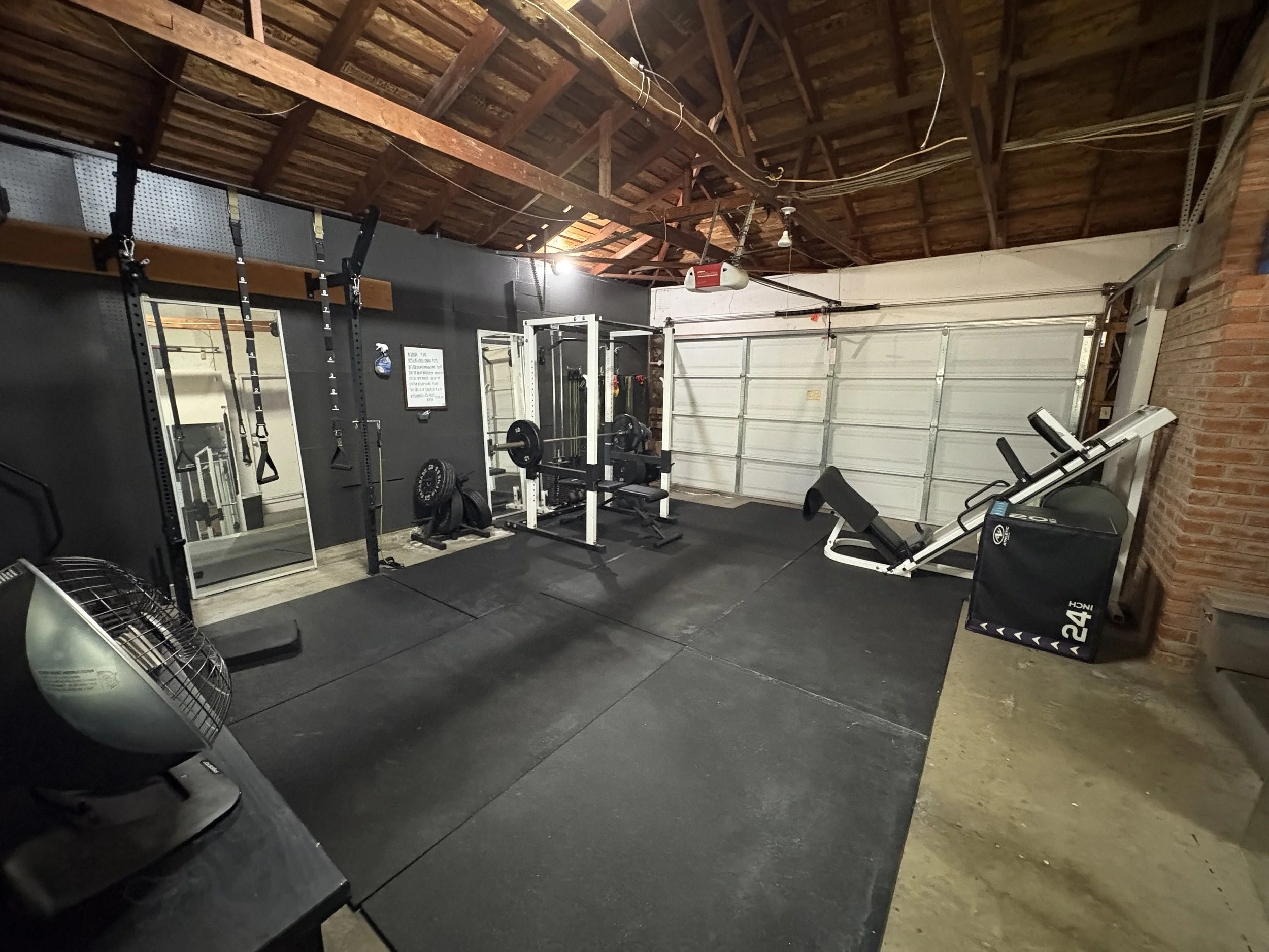 A home gym with various workout equipment including pull-up bars, a mirror, barbells, a squat rack, an exercise bench, a plyometric box, and a fan. The space has a black rubber floor, exposed wooden ceiling, and a garage door on the back wall.
