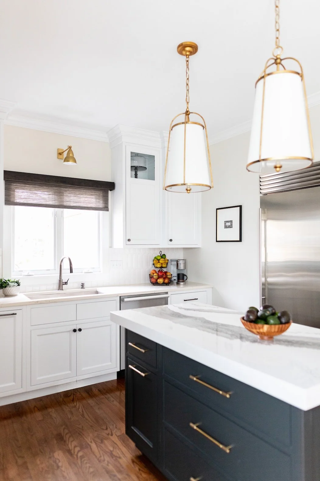 Modern kitchen with white cabinets, a black island with gold handles, a white marble countertop, and two hanging pendant lights with white shades and gold accents. There's a window with a brown blind, a bowl of avocados on the island, and fruits on a tiered stand on the countertop.