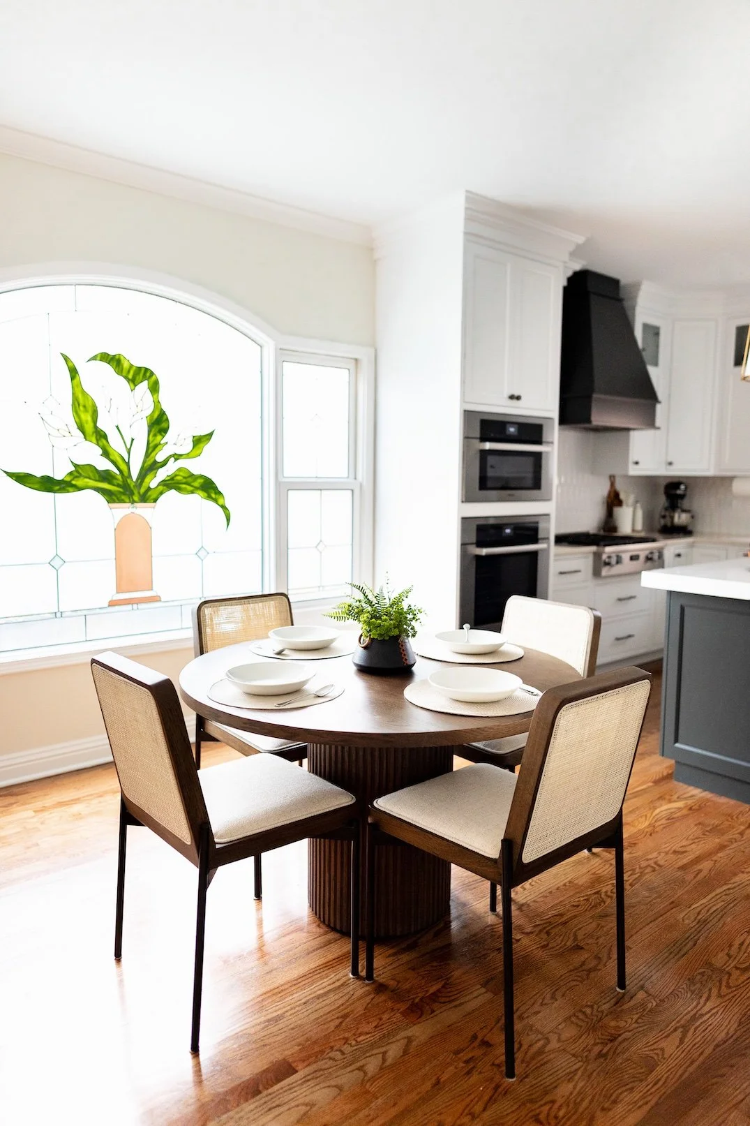 A dining area in a bright kitchen with a round wooden table set with white plates and bowls, four upholstered chairs, a potted plant in the center, and a large decorative window with a stained glass pattern of a potted plant.