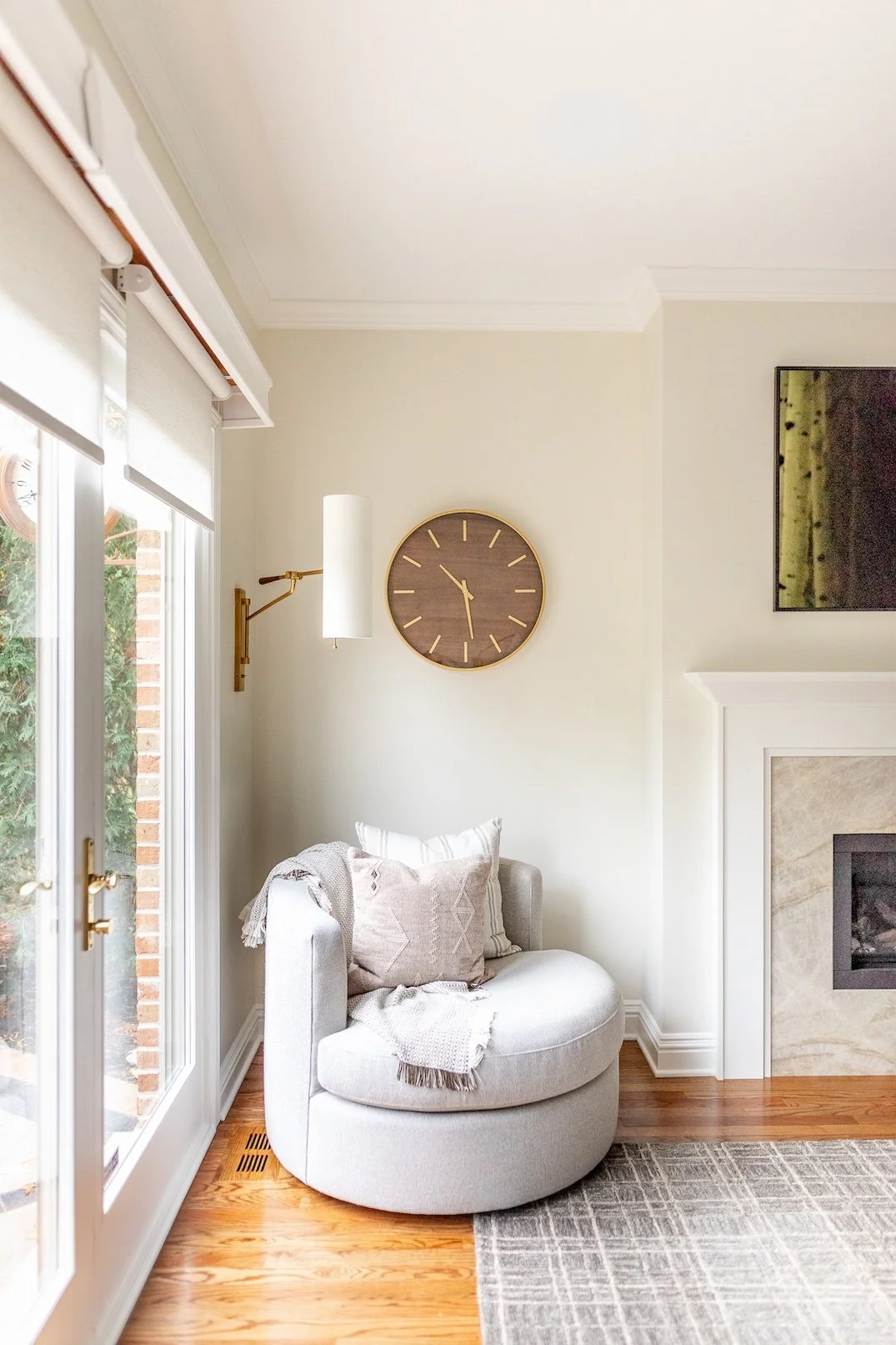 A cozy corner with a light gray circular chair, decorative pillows, a blanket, near large windows with blinds, a wall-mounted clock, a modern fireplace, and a flat-screen TV on the wall.