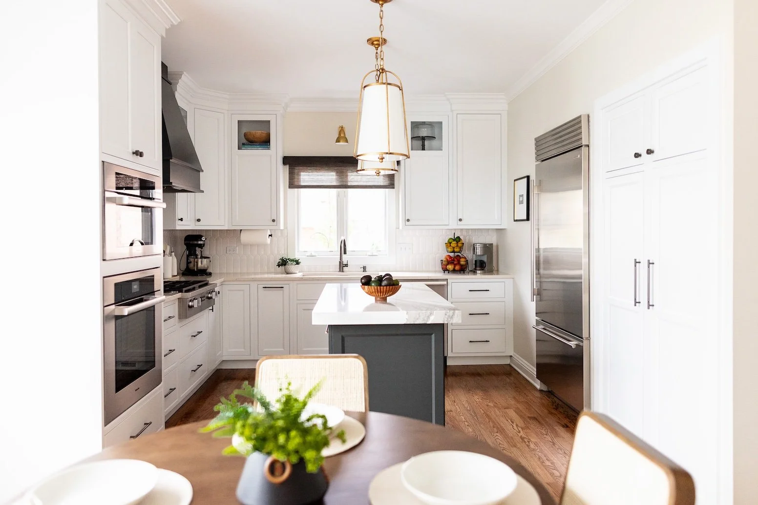 Modern kitchen with white cabinets, stainless steel refrigerator, and a central island with a white marble top. There are wooden floors, a window with blinds, and a hanging light fixture over the island. A dining table with a plant centerpiece is in the foreground.