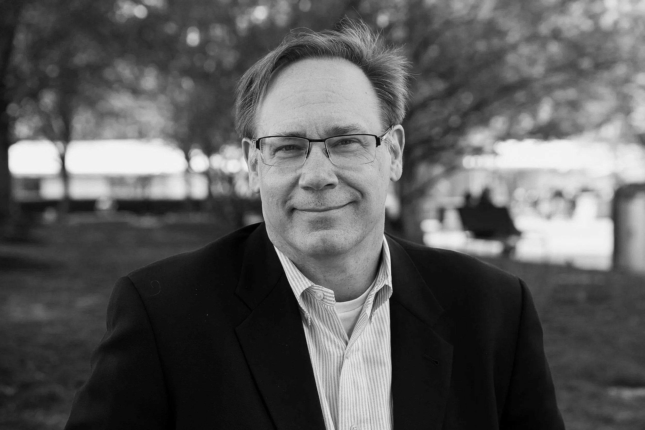 Black and white photo of a man in glasses, a collared shirt, and a blazer, smiling outdoors with trees and benches in the background.