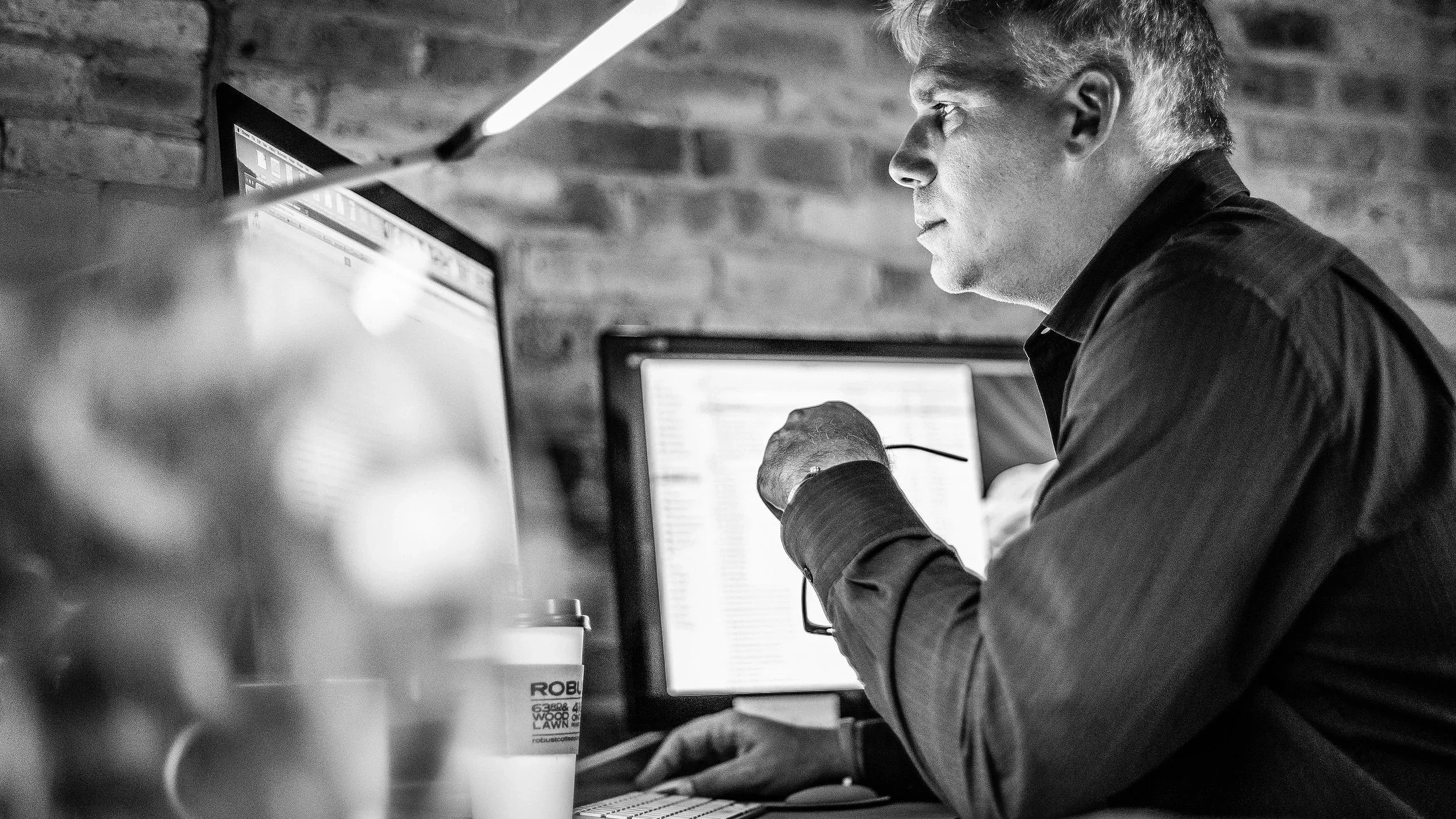 A man working at a computer in a modern workspace with brick walls, wearing a dark shirt, focused on his screen.