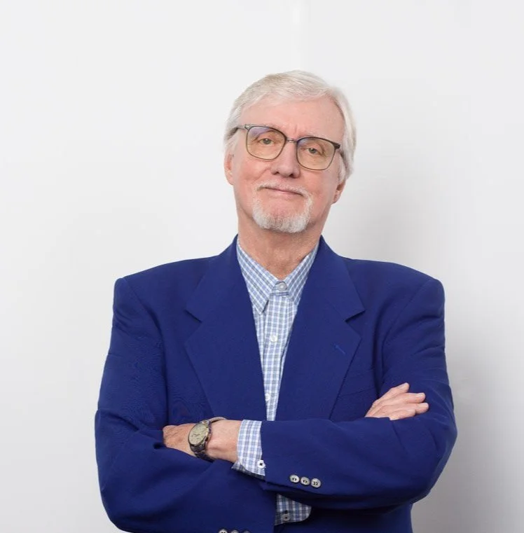 A senior man with gray hair and glasses, wearing a blue blazer and a checkered shirt, standing with arms crossed against a plain white background.