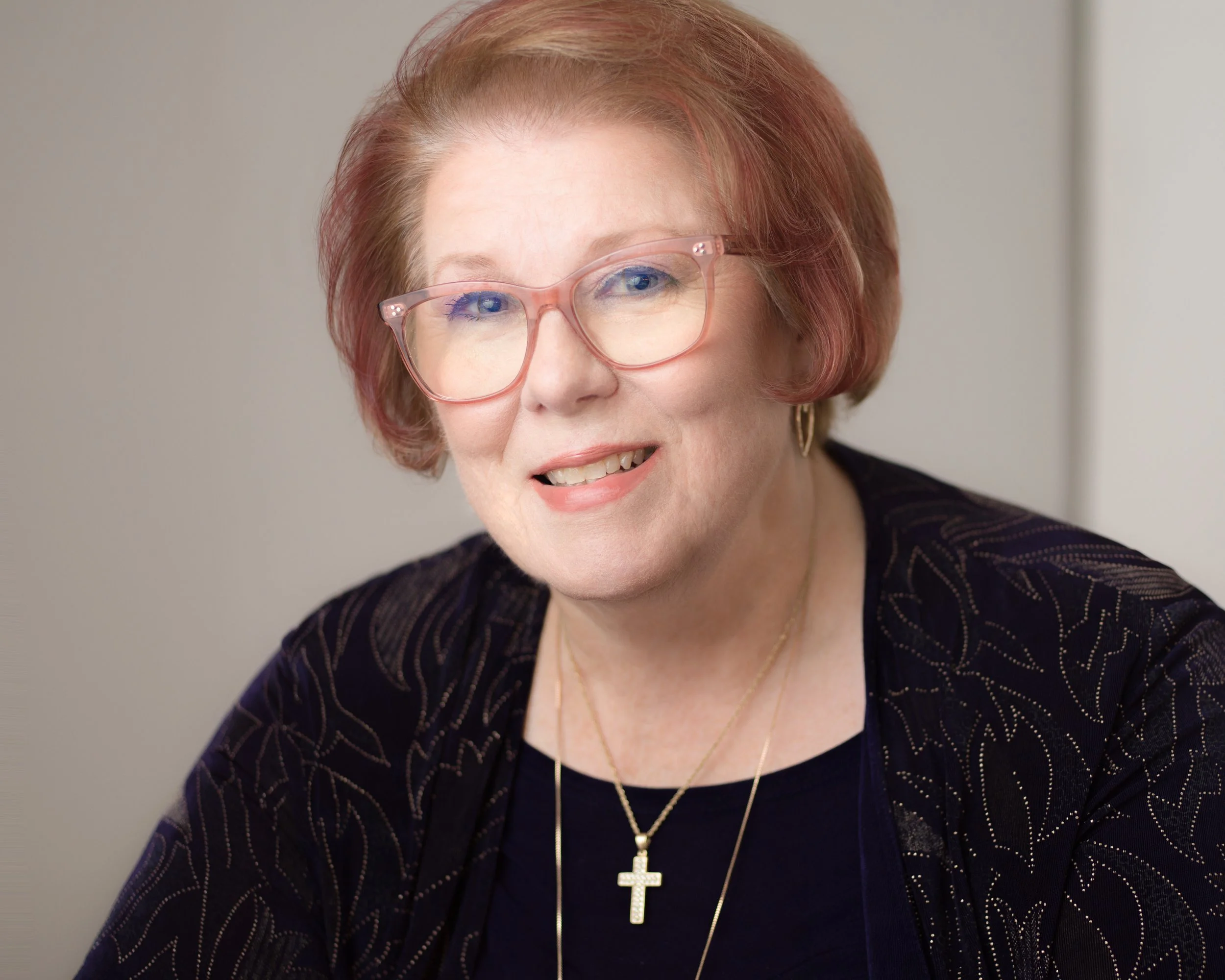 A smiling elderly woman with short, reddish hair, wearing pink glasses, gold earrings, and a gold cross necklace, dressed in black with a dark blazer that has a dotted line pattern.
