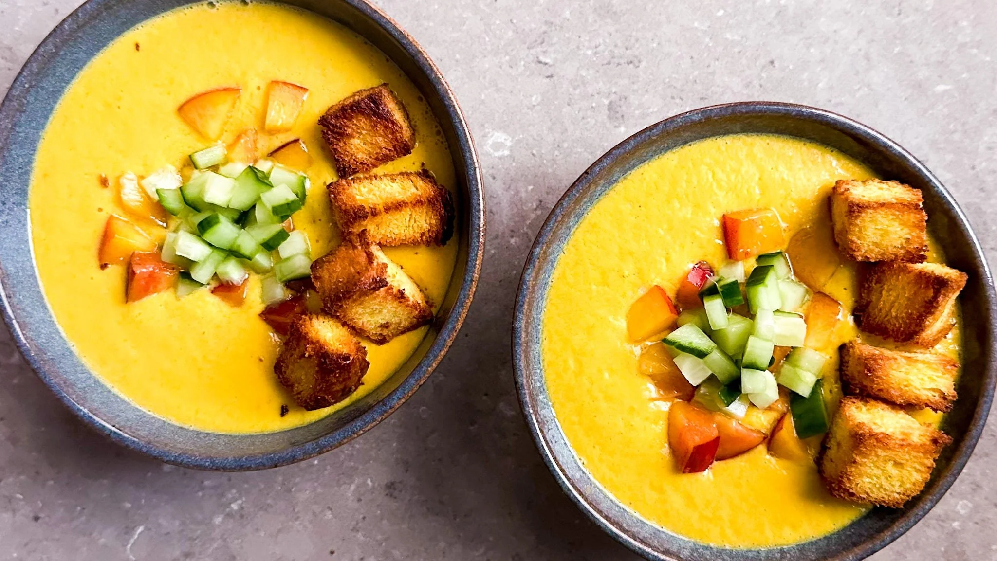 Photo of yellow tomato gaspacho with garnishes of peaches cucumber and croutons.