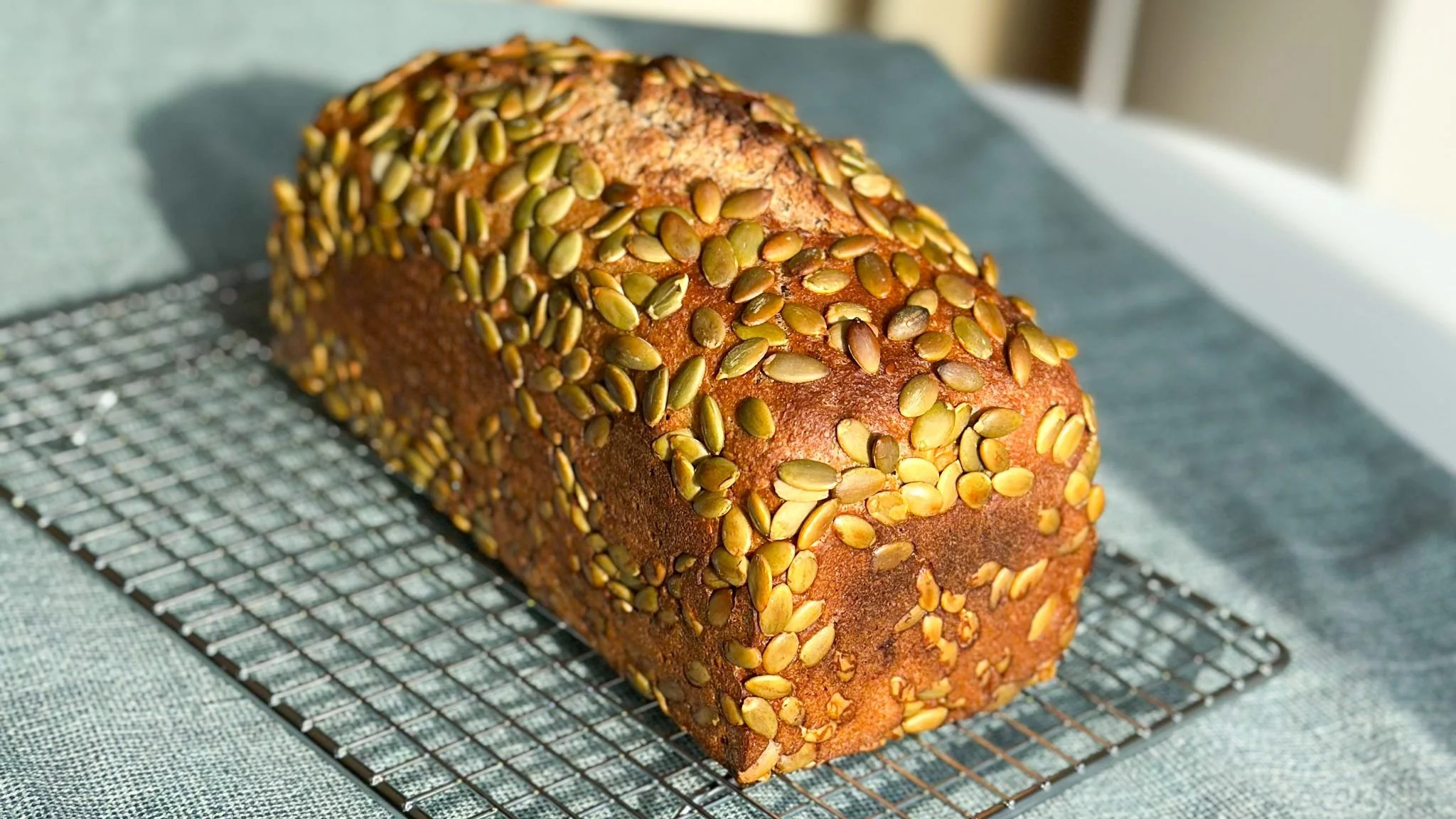 Photo of a loaf of whole wheat bread, covered in pumpkin seeds, on a wire cooling rack.