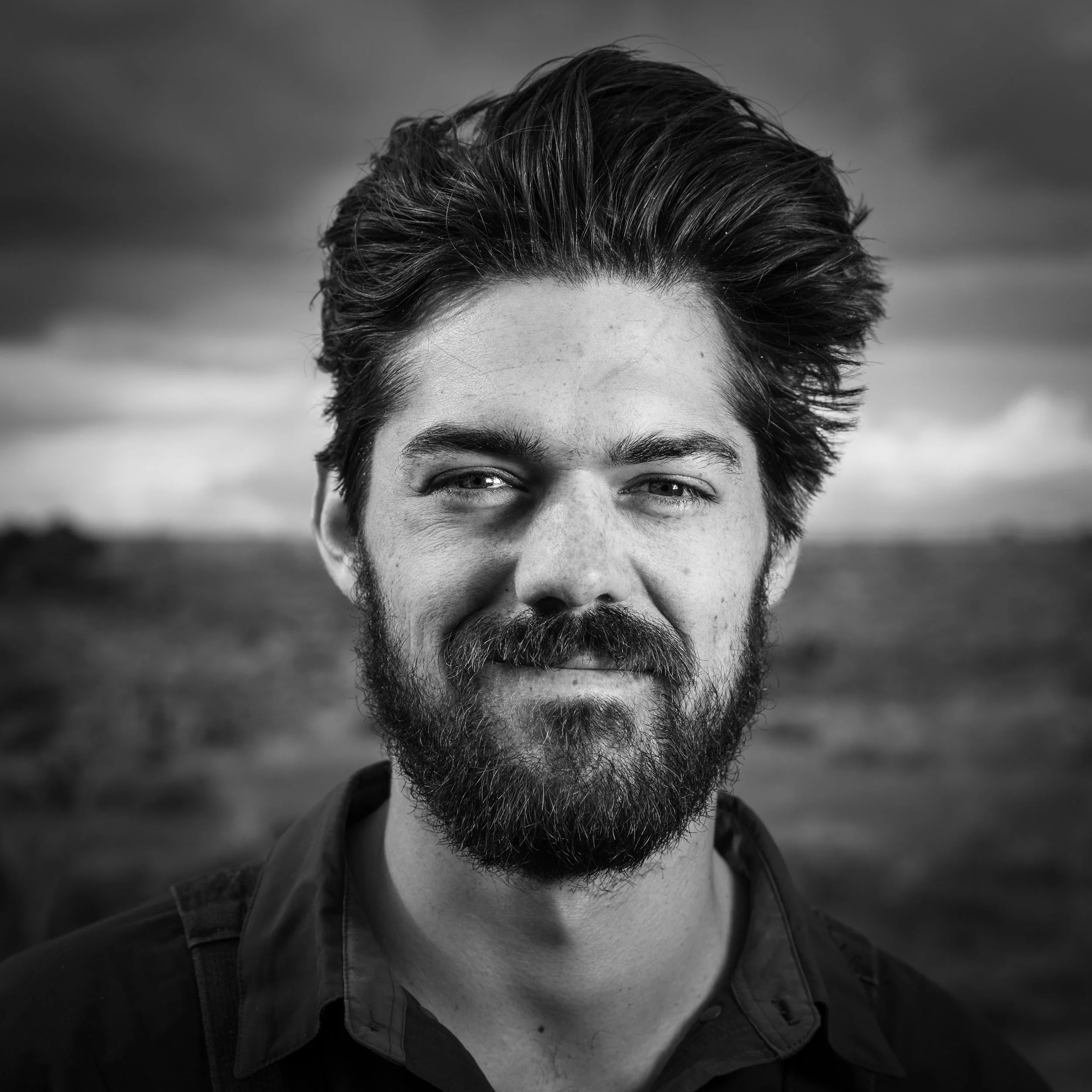 Black and white portrait of a smiling man with a beard and thick hair outdoors.