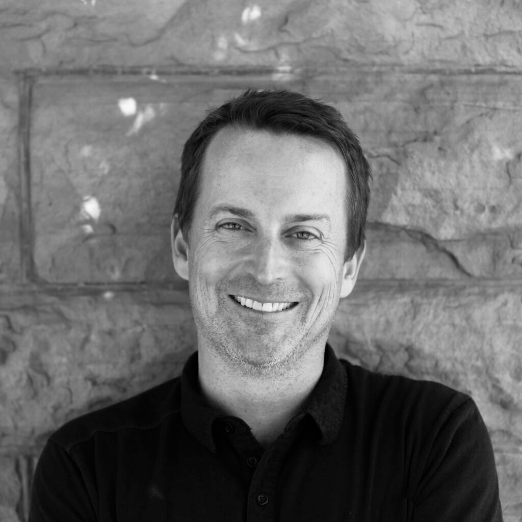 Black-and-white portrait of a smiling man with short hair, wearing a dark collared shirt, standing against a stone wall.