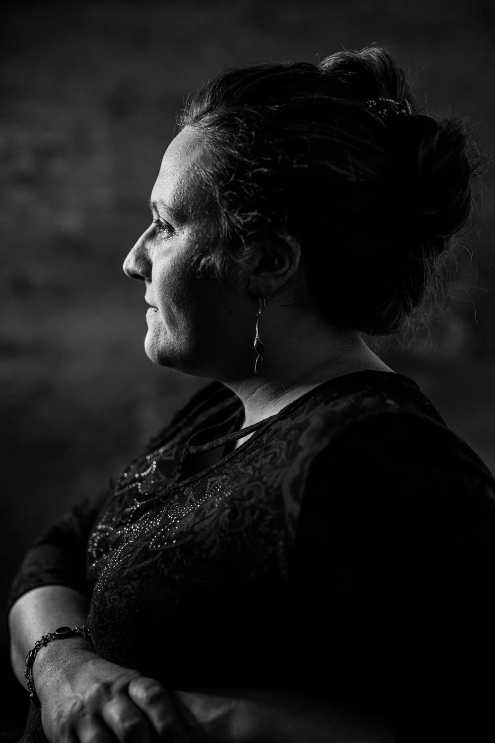 Black and white side profile of a middle-aged woman with styled hair, wearing jewelry and an ornate top, against a dark background.