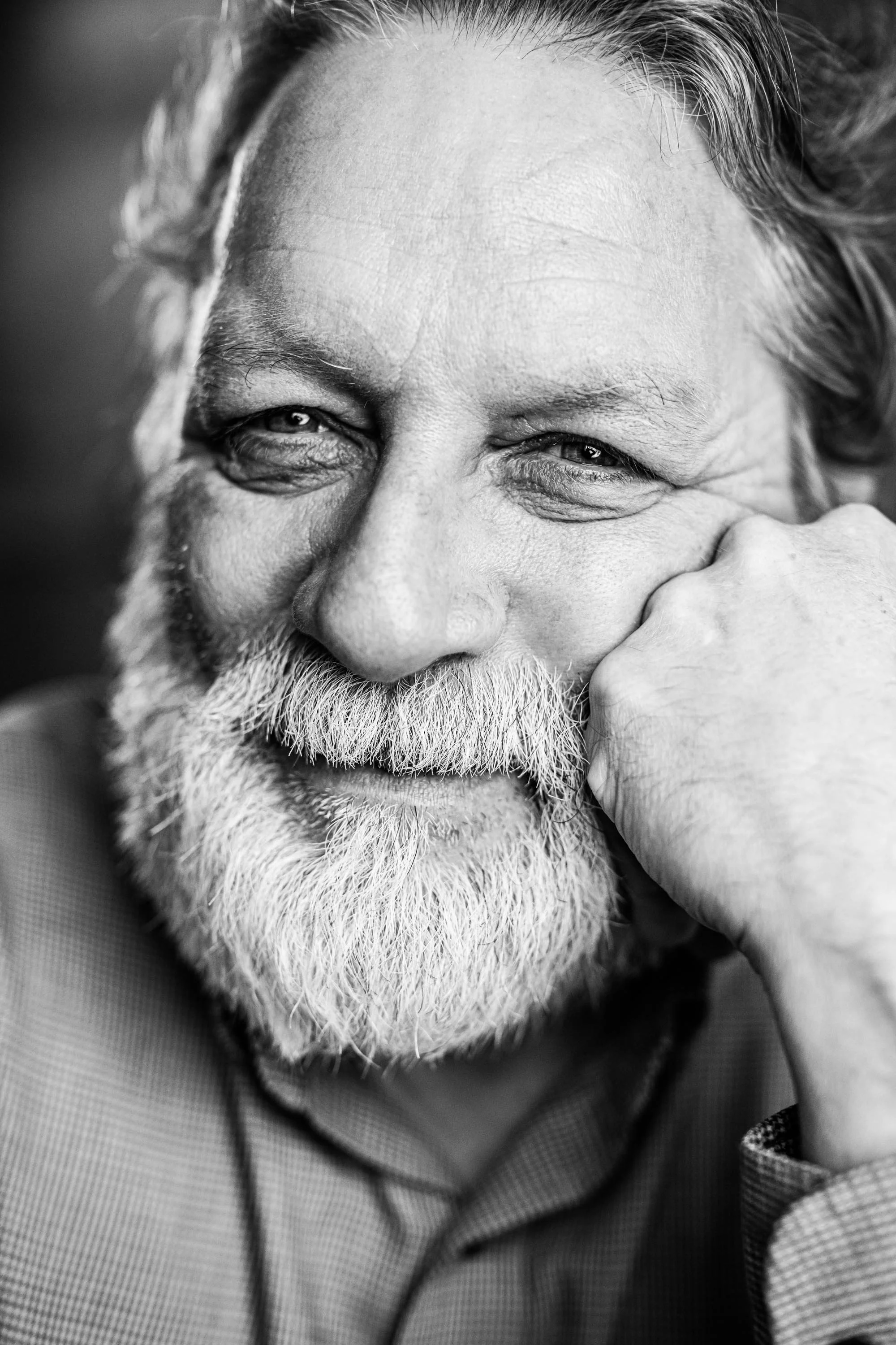 Close-up black and white photo of an elderly man with a beard, smiling gently, resting his face on his hand.
